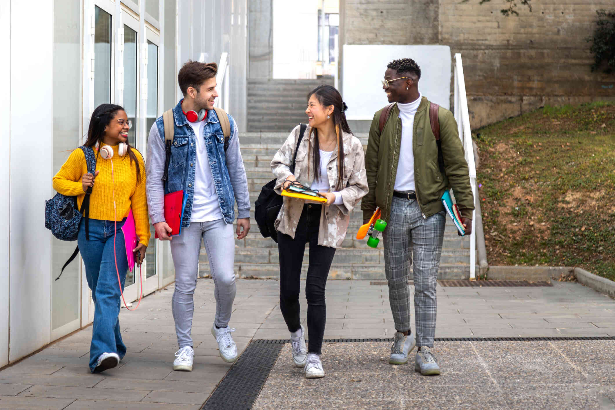 A group of teens walk down the steps of a school campus while holding books and backpacks and engaging in a conversation with positive expressions