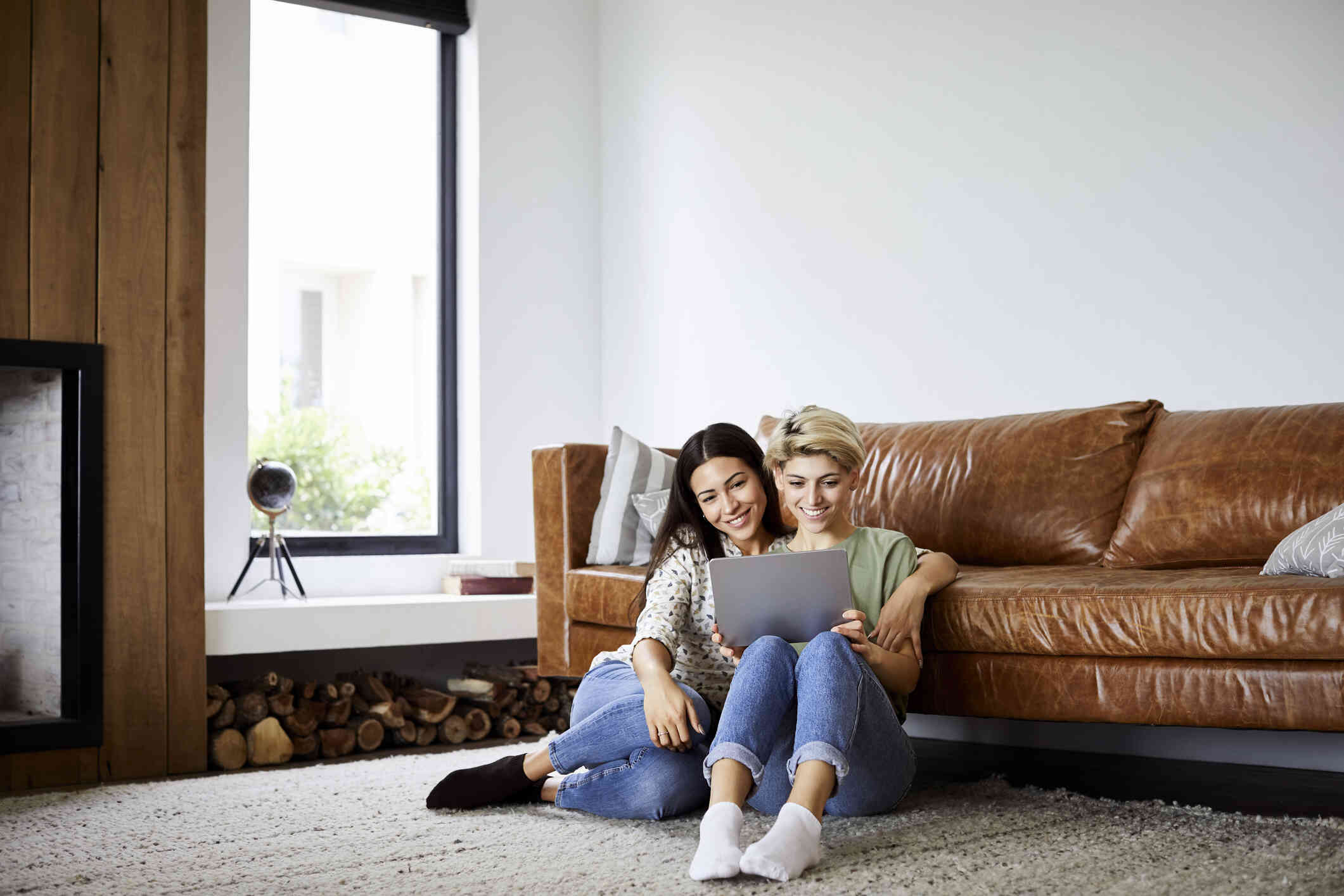A female couple sit close together on the livingroom floor with their backs against the couch and look at the laptop in their lap with a smile.