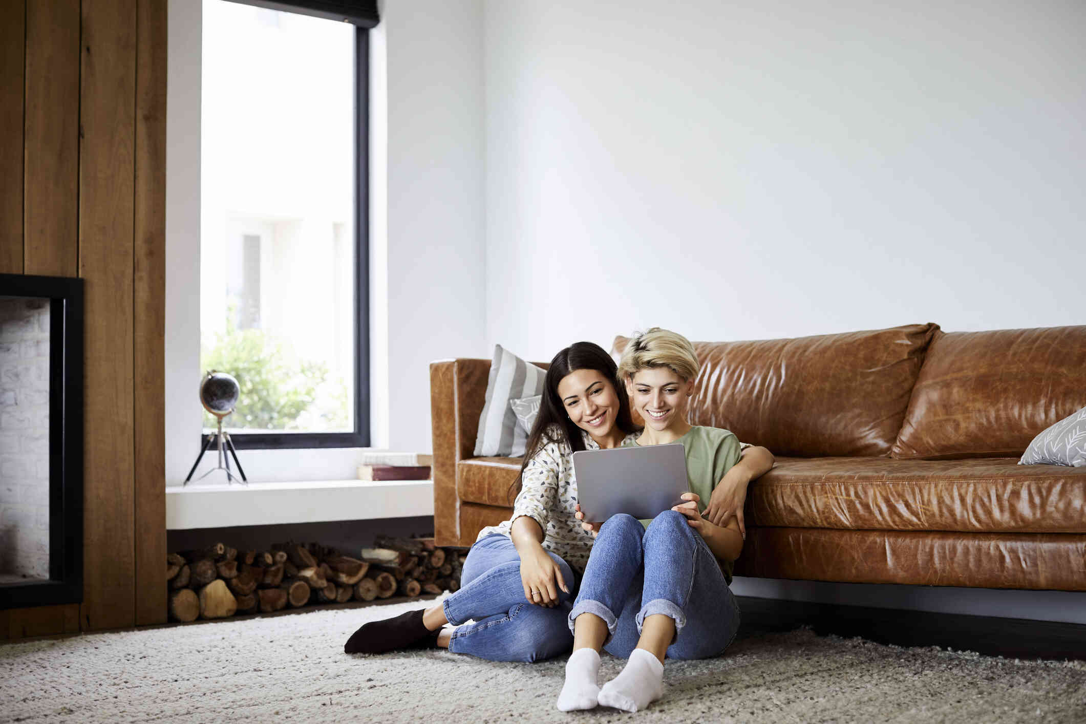 A female couple cuddle on the living room floor while looking at a laptop and smiling.