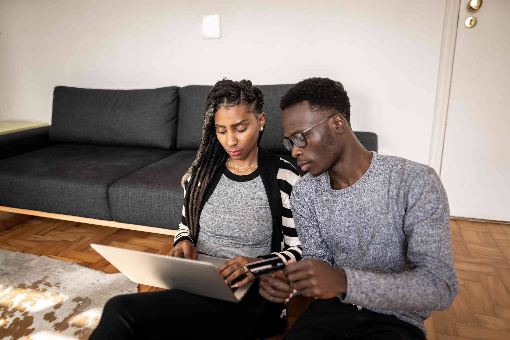 A woman with braids looks down at her computer with a serious expression while a man with glasses sits next to her on the floor.