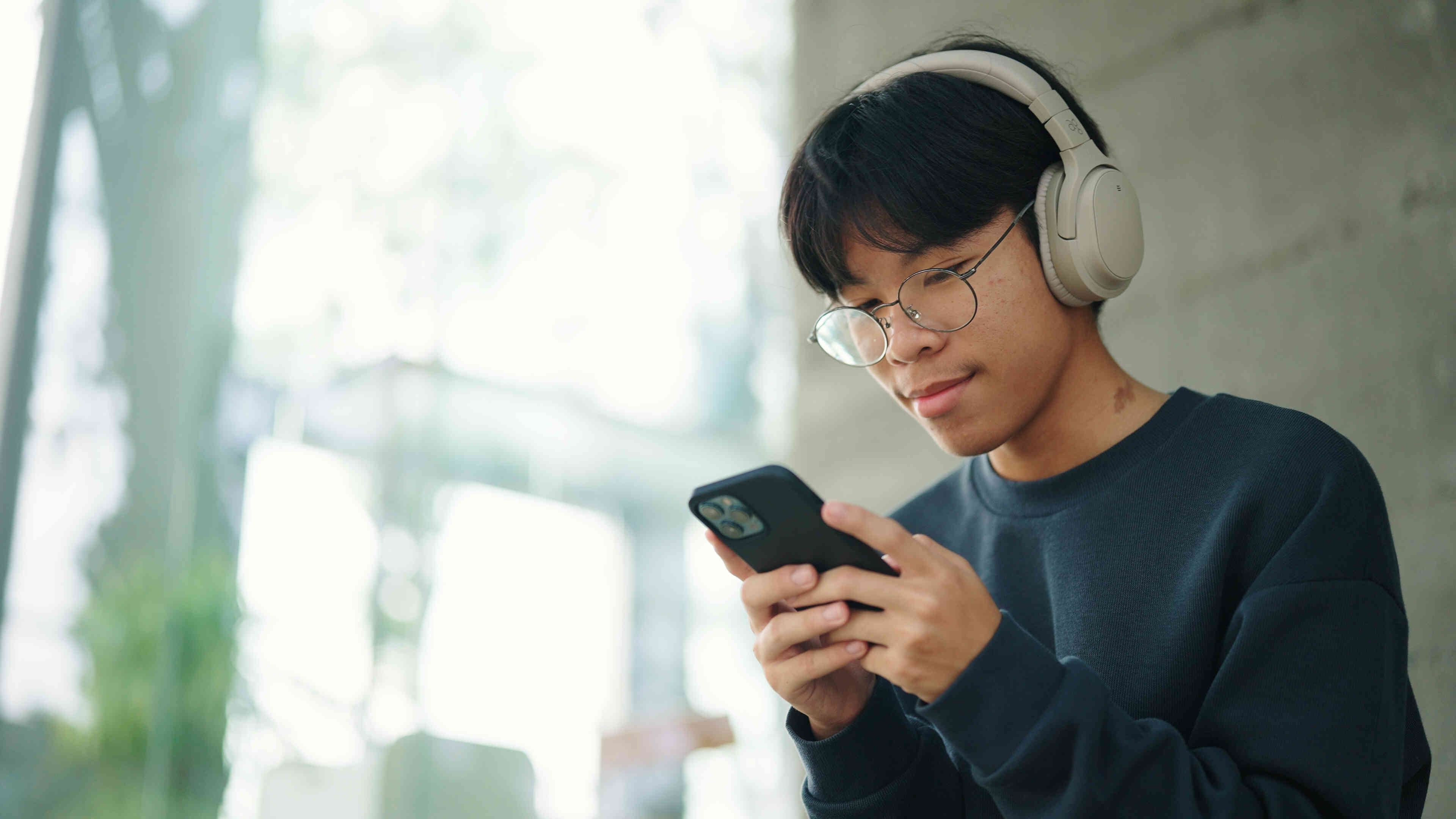 A close up of a teen boy wearing headphones as he looks down at the cellphone in his hands.