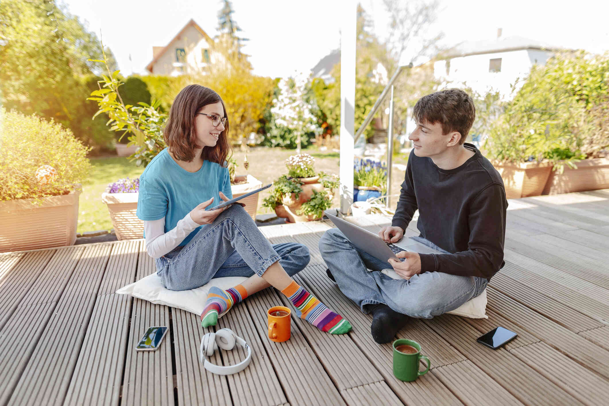 A young man sits outside on a porch with a laptop and a coffee mug as a young woman wearing glasses sits next to him with a tablet, headphones, and a coffee mug.