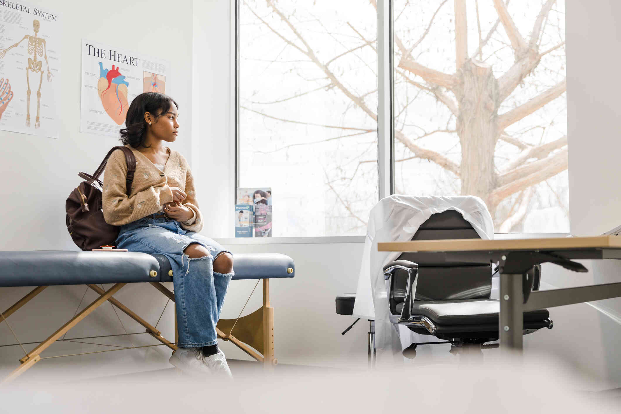 A woman with a backpack sits on a medical table in a doctors office while gazing off with a worried expression.