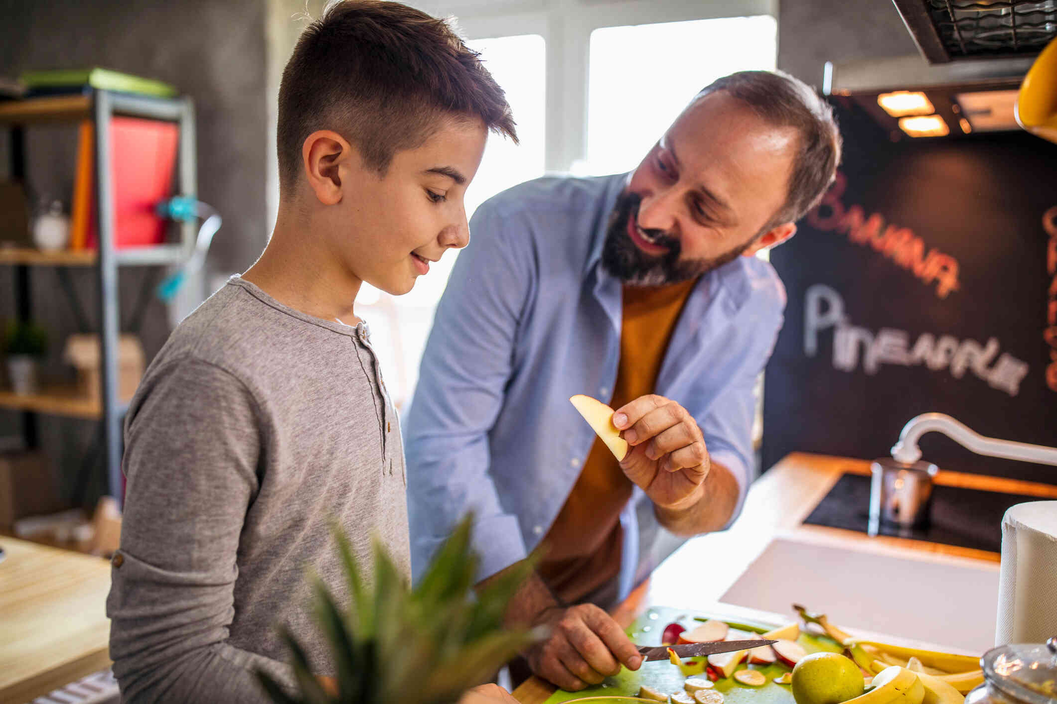 A man with a beard smiles as he holds a slice of apple up to a young boy standing next to him in a kitchen cutting fruit.