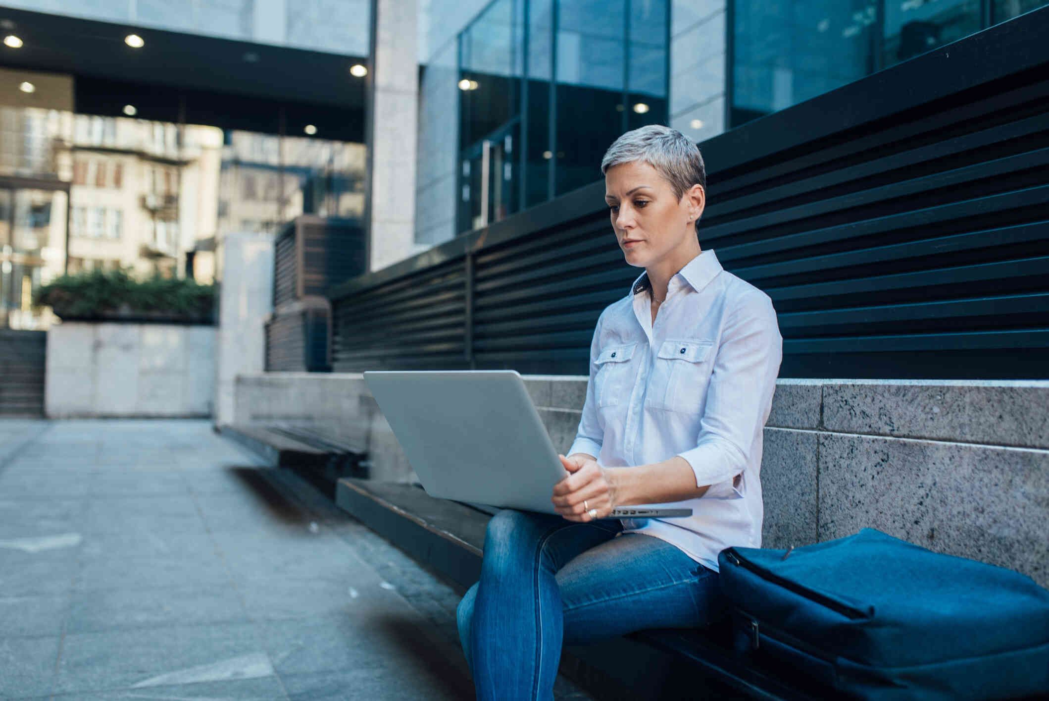 A woman in a white shirt sits on a bench outside of a  large building and types on the laptop in her lap.