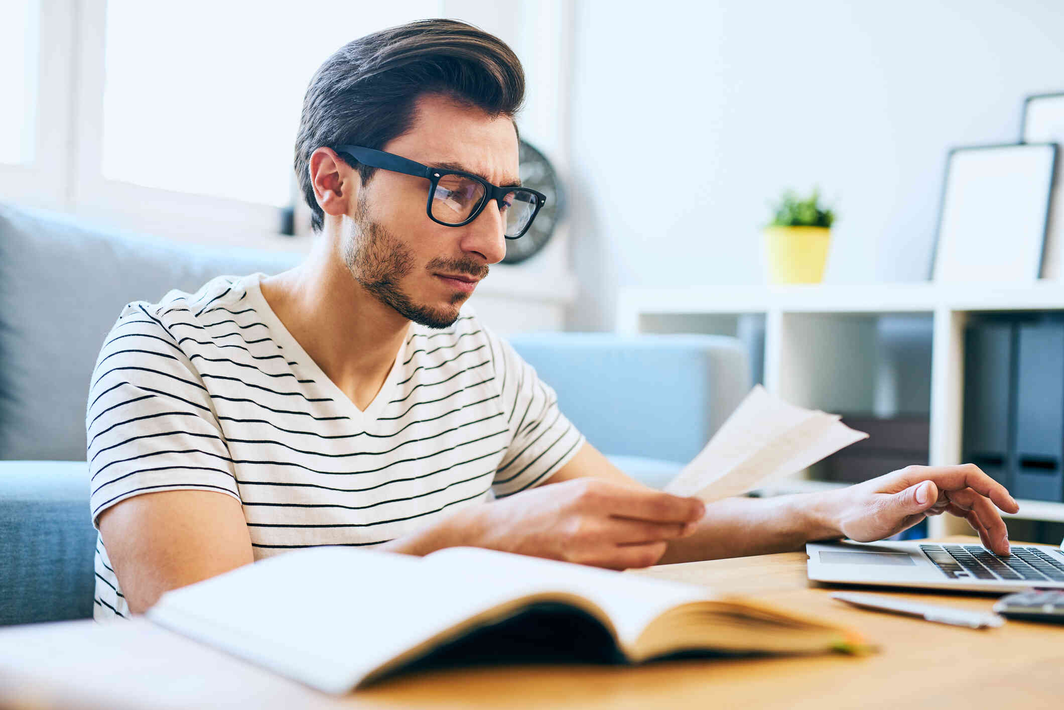 A man with glasses sits at a table with a book open next to him as he looks at some papers in his hand and types on his laptop.