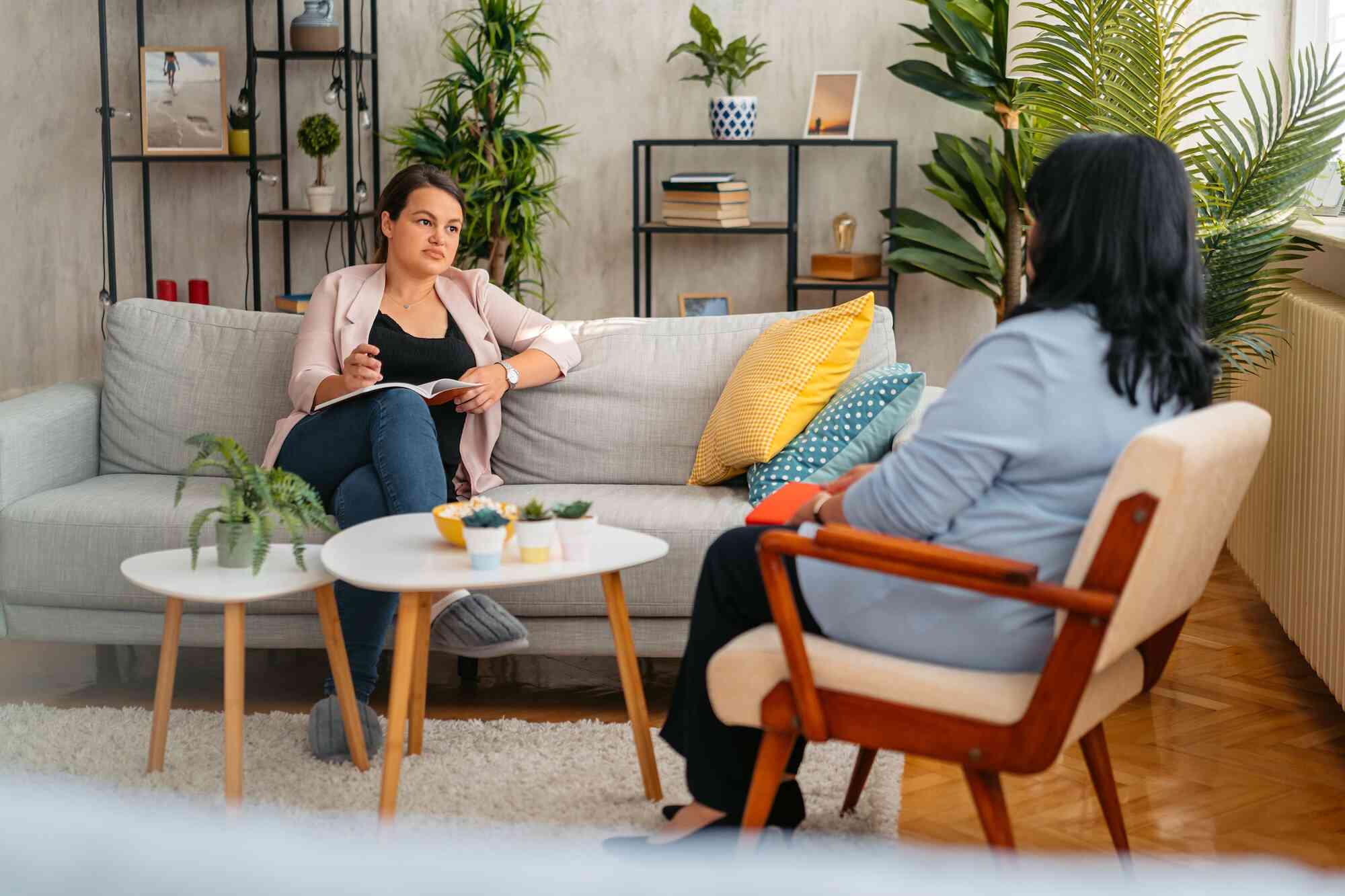 A woman in a blue shirt sits in a chair while talking to a woman on the couch across. 