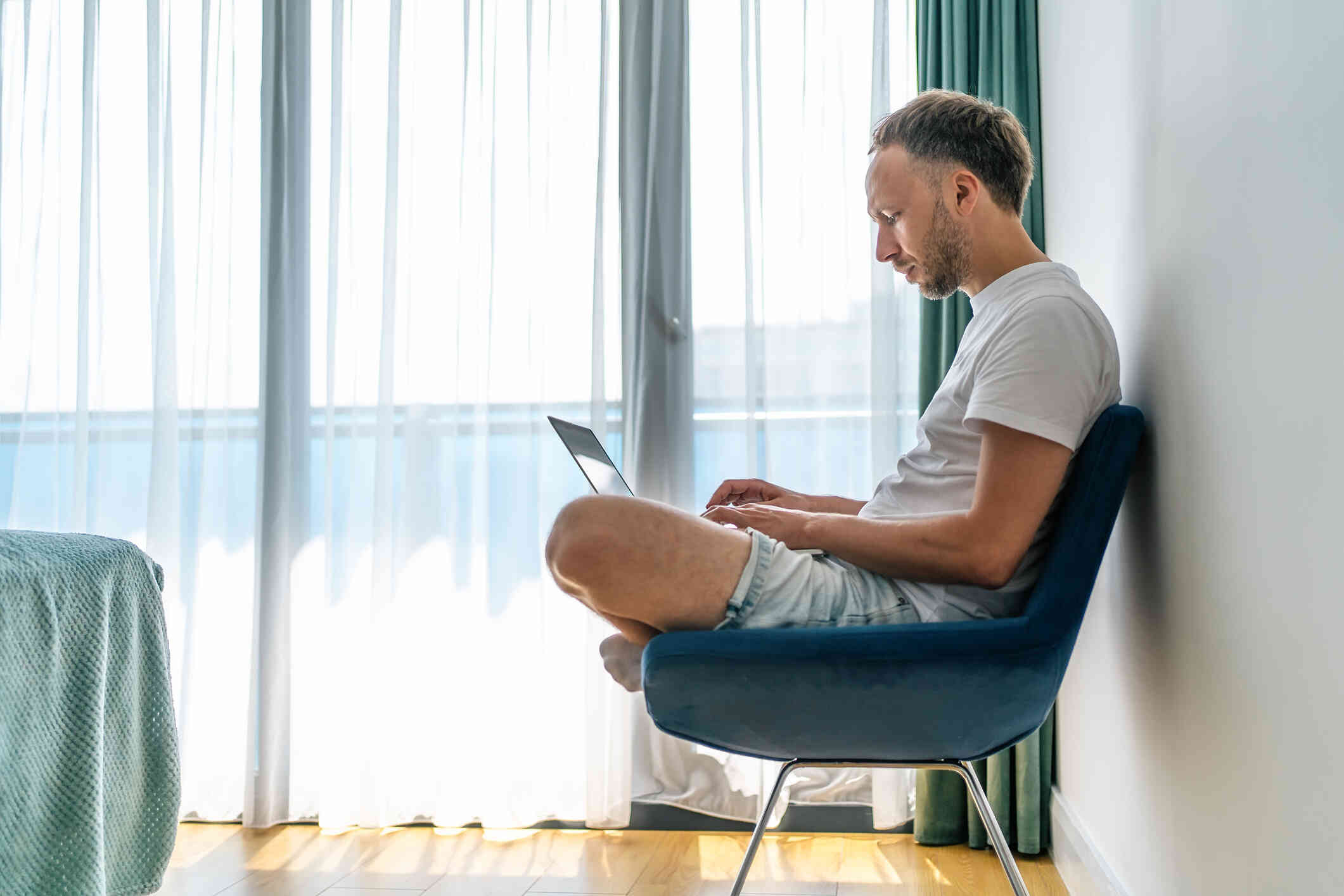 A man sits cross legged in a chair near a window and looks down at the laptop in his lap.