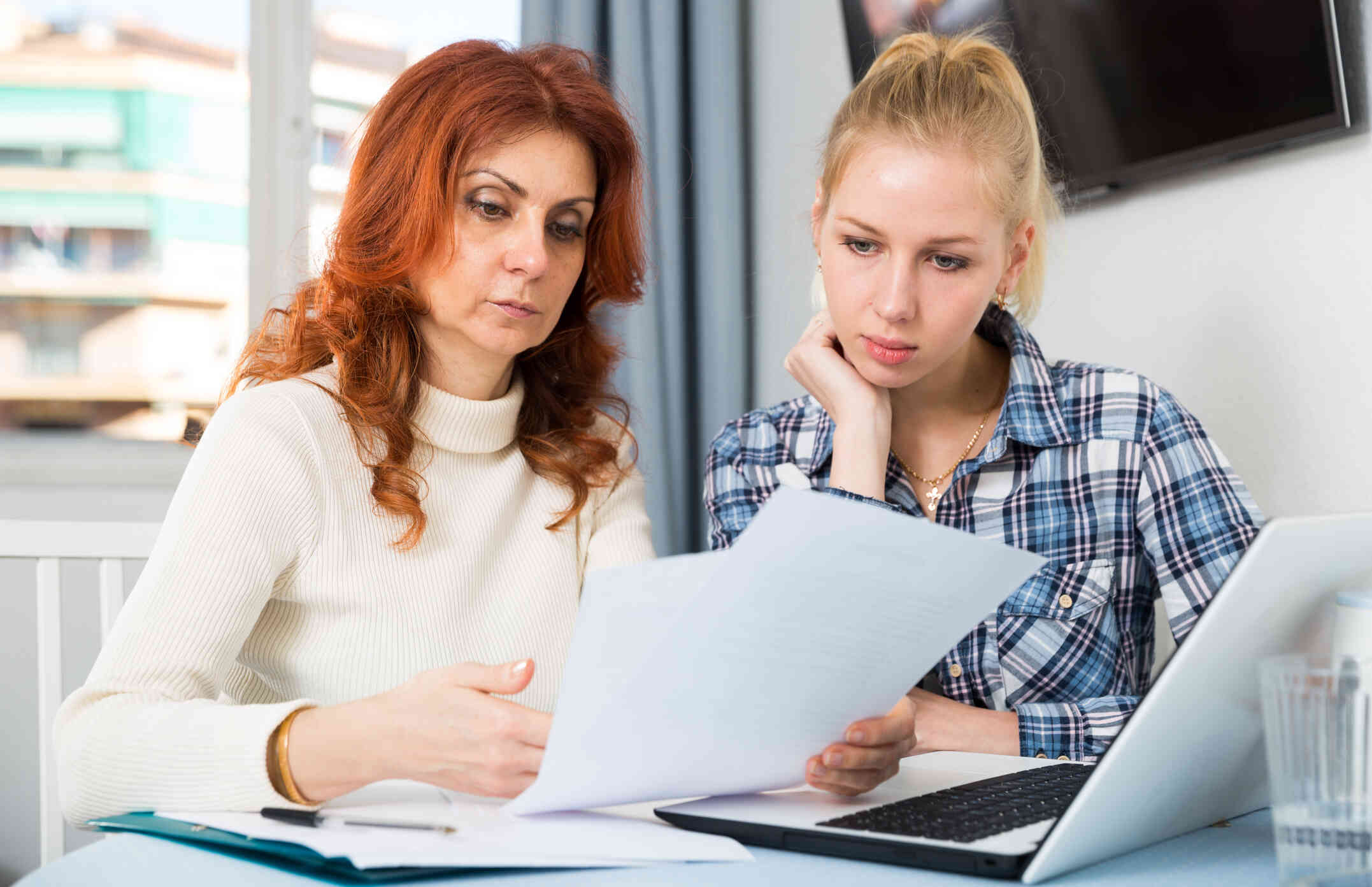 A woman with a serious expression and red hair looks down at a piece of paper she is holding in her hand. Another woman with blonde hair sits next to her and looks down at the paper.