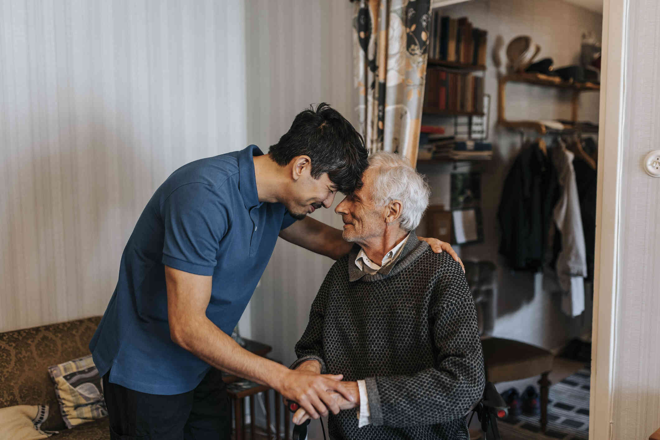 An elderly man sits on his walker in his home while a younger male family member leans forwards and gently rests his forehead against the older mans as they both smile softly.