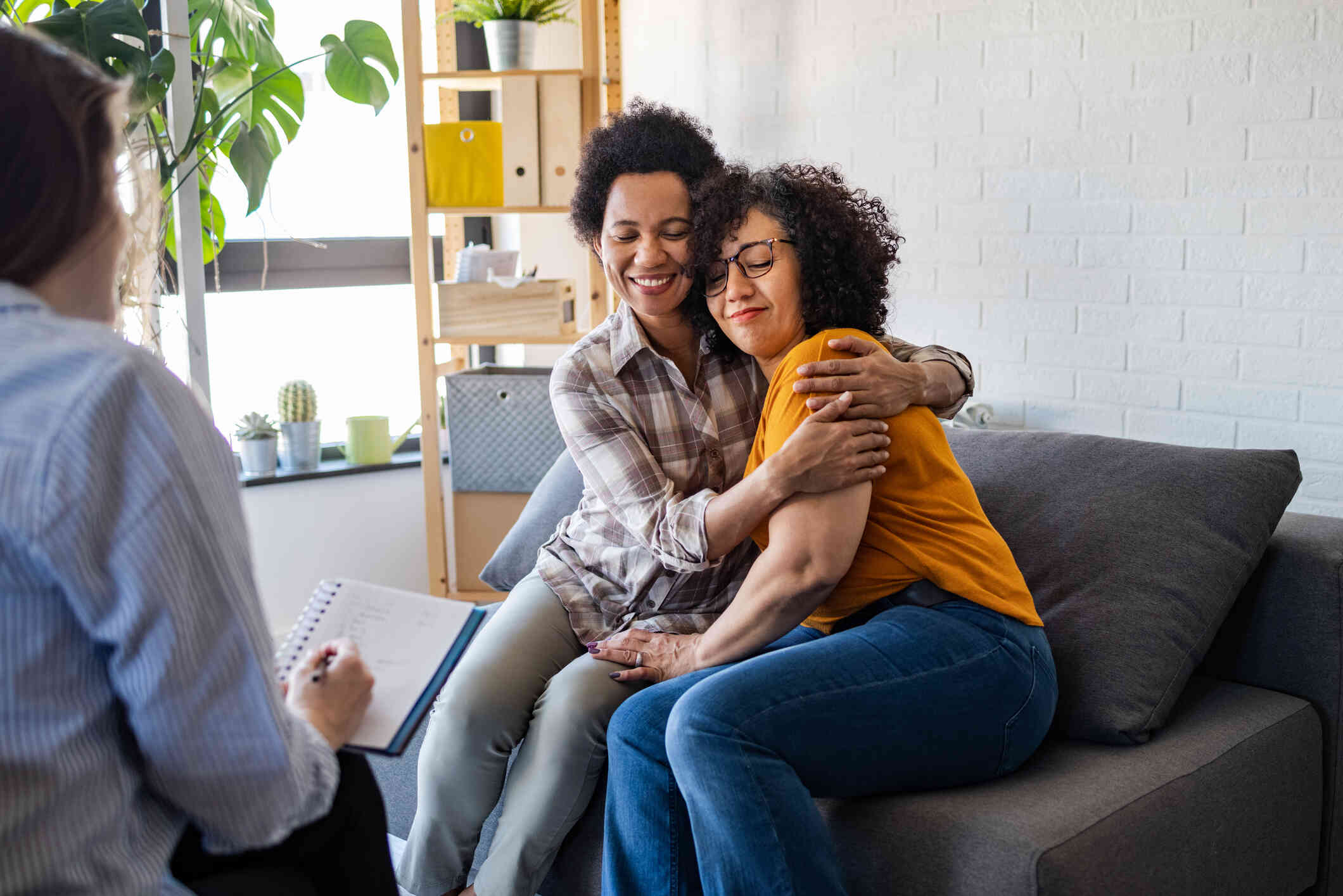 Two women embrace lovingly while sitting on a couch across from their therapist during a therapy session.