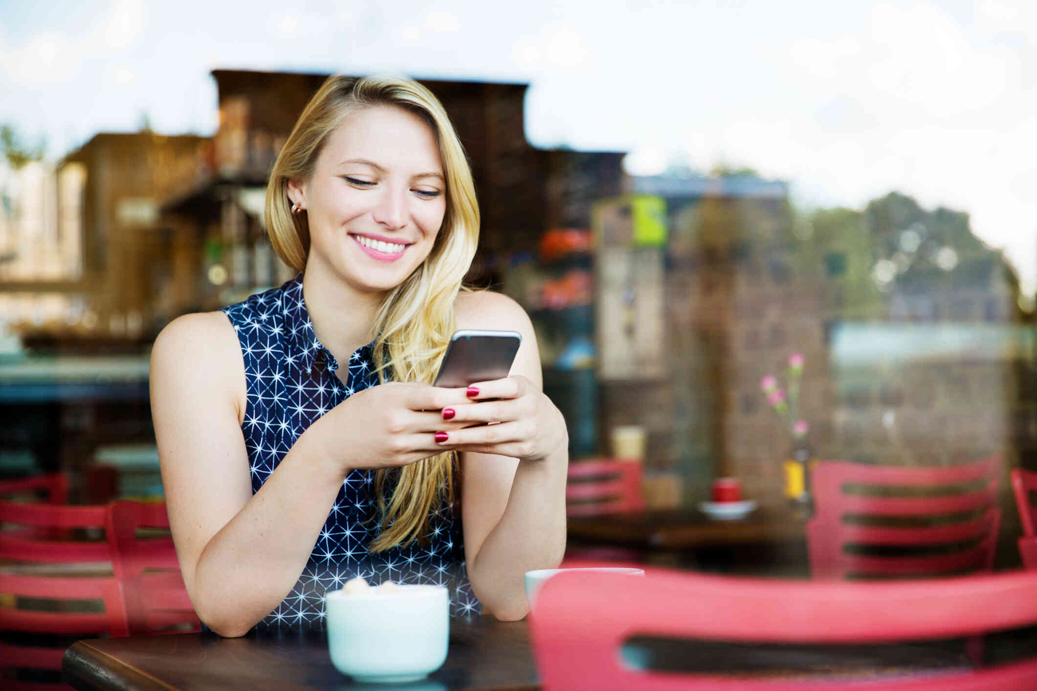 A woman in a blue shirt sits at a cafe table and smiles while looking down at the phone in her hand.