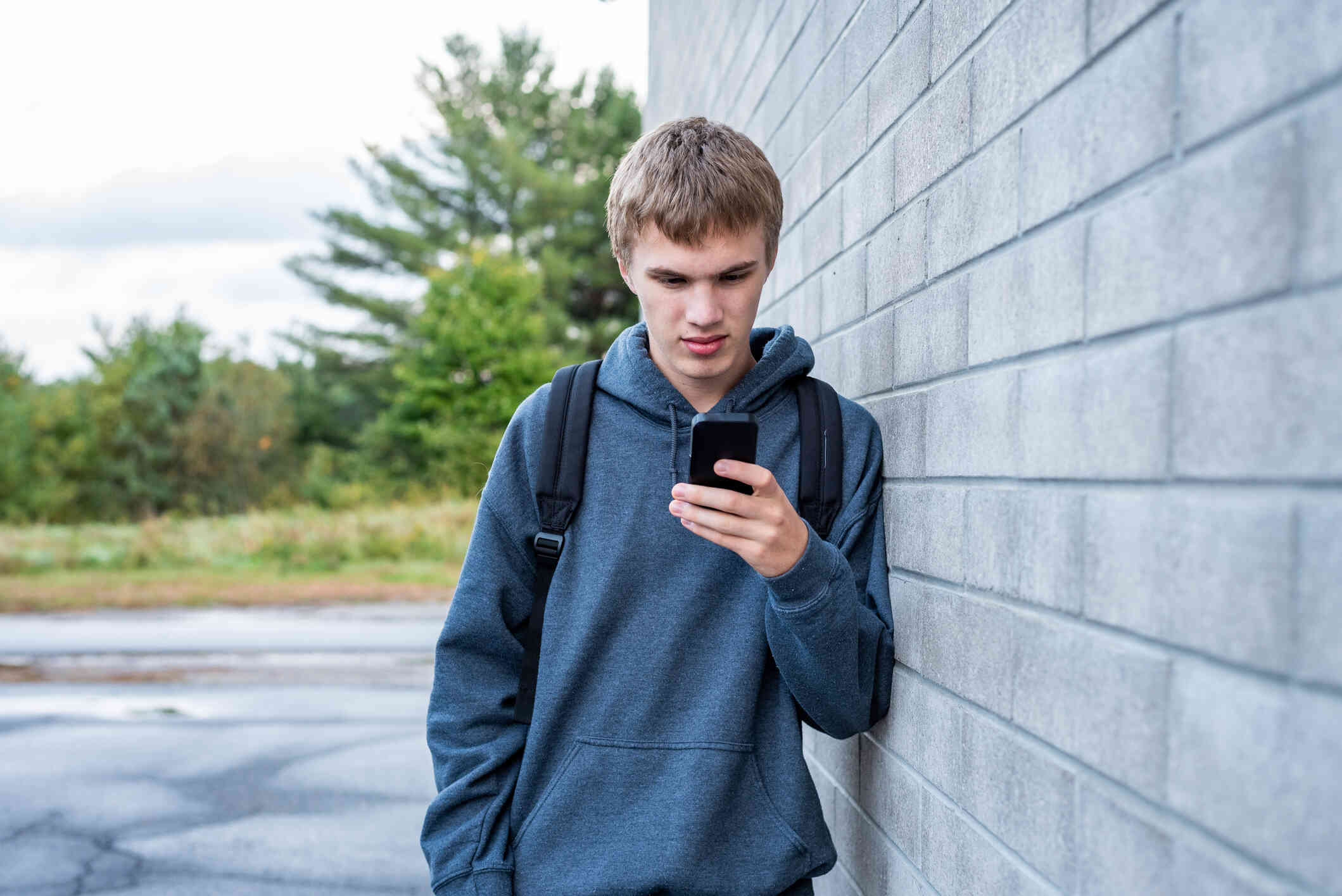 A man in a sweater and backpack leans his shoulder against a brick wall while looking down at his phone with a worried expression.