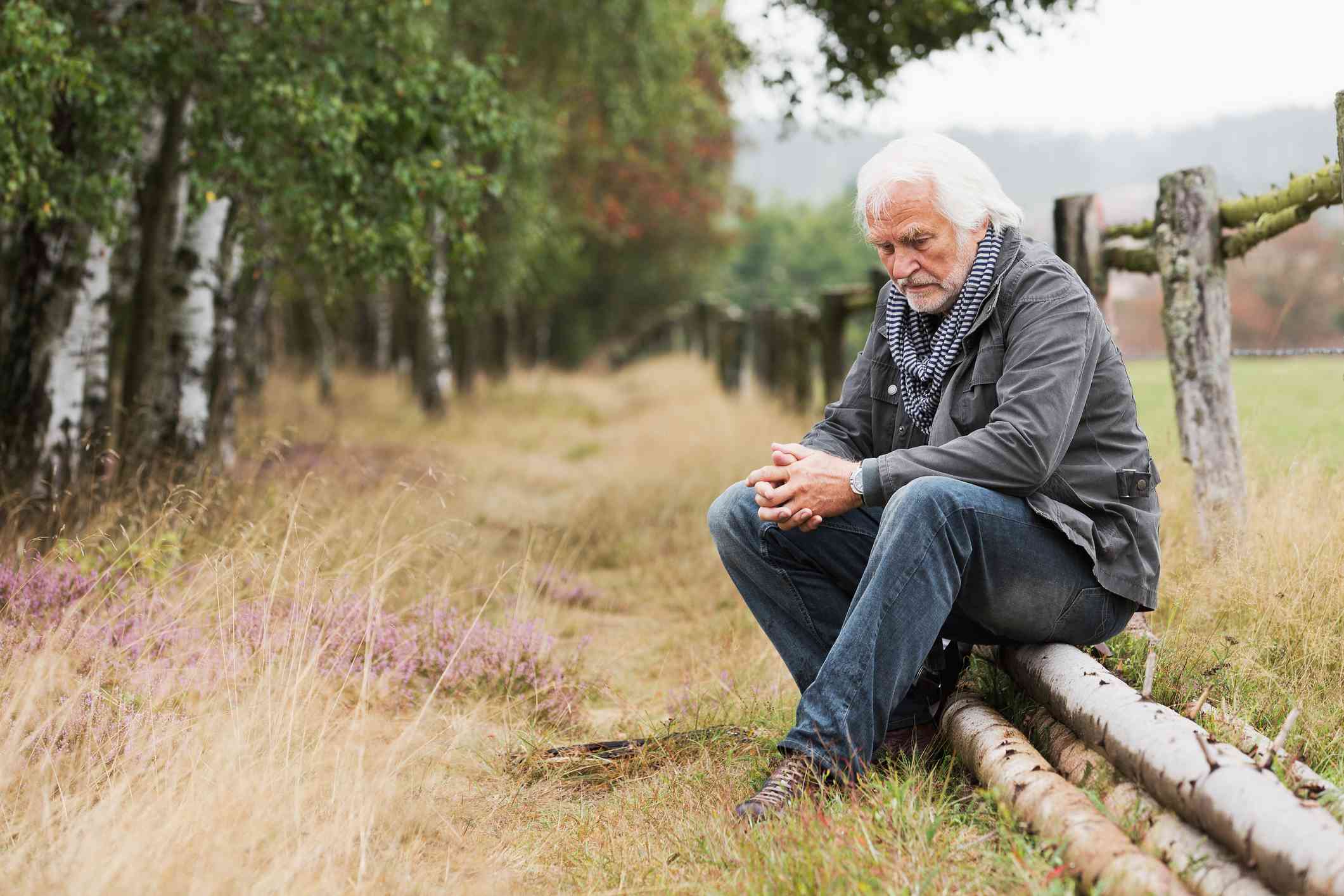 An elderly man sits on some fallen trees outside on a cloudy day with his hands clasped together deep in thought.