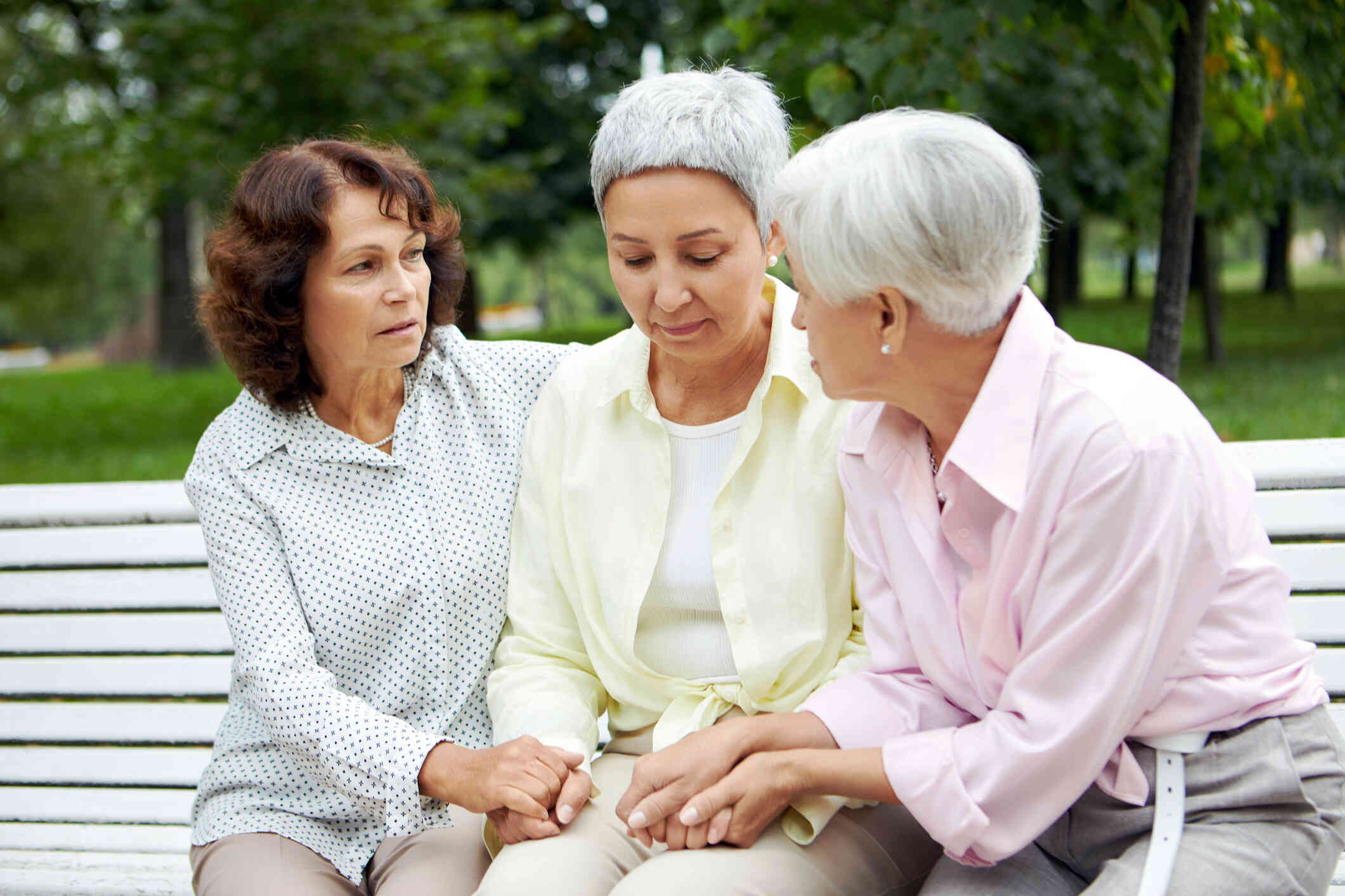 Three elderly woman sit on a park bech outside on a sunny day with sad expressions.