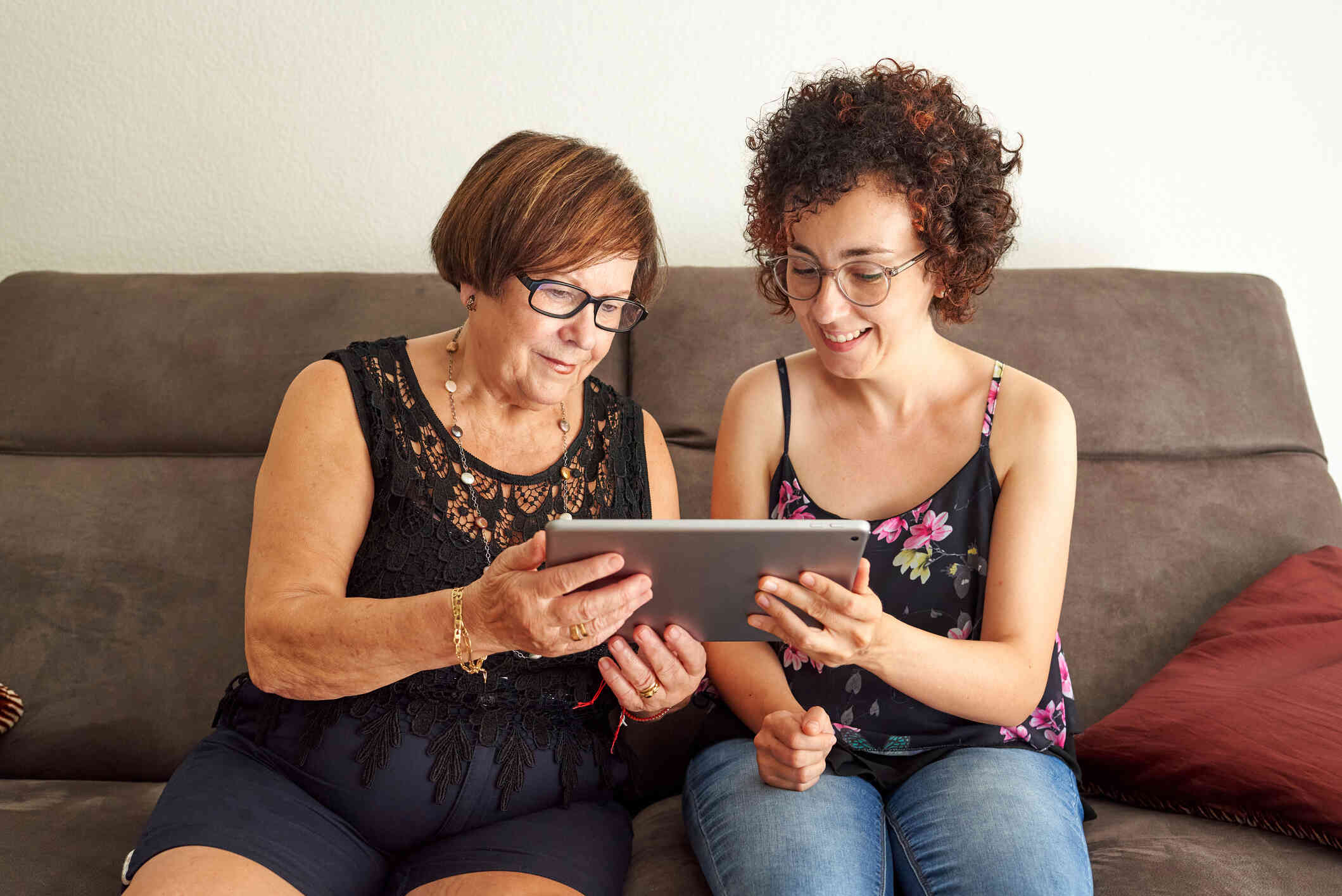 A middle aged woman and her younger female relative sit next to each other on the couch while looking at a tablet together and smiling.