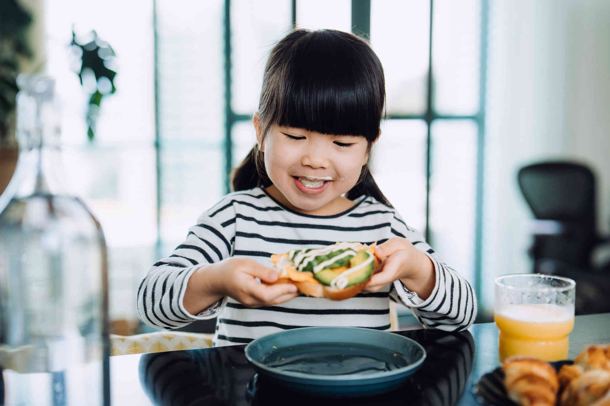 A little girl sits at the kitchen table and eats on open face sandwhich while smiling.
