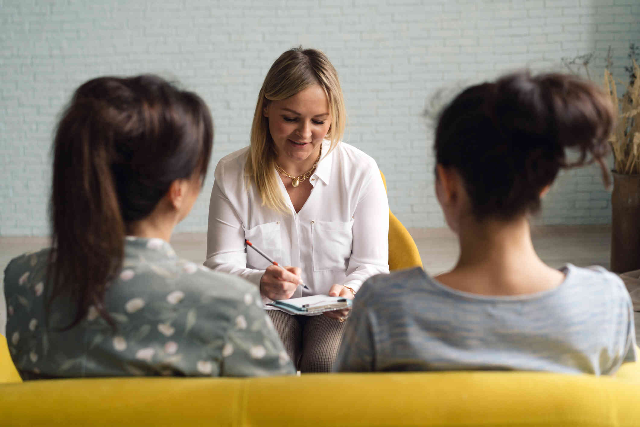 Two female family members sit next to each other while the female therapist across from them writes on her clipboard.