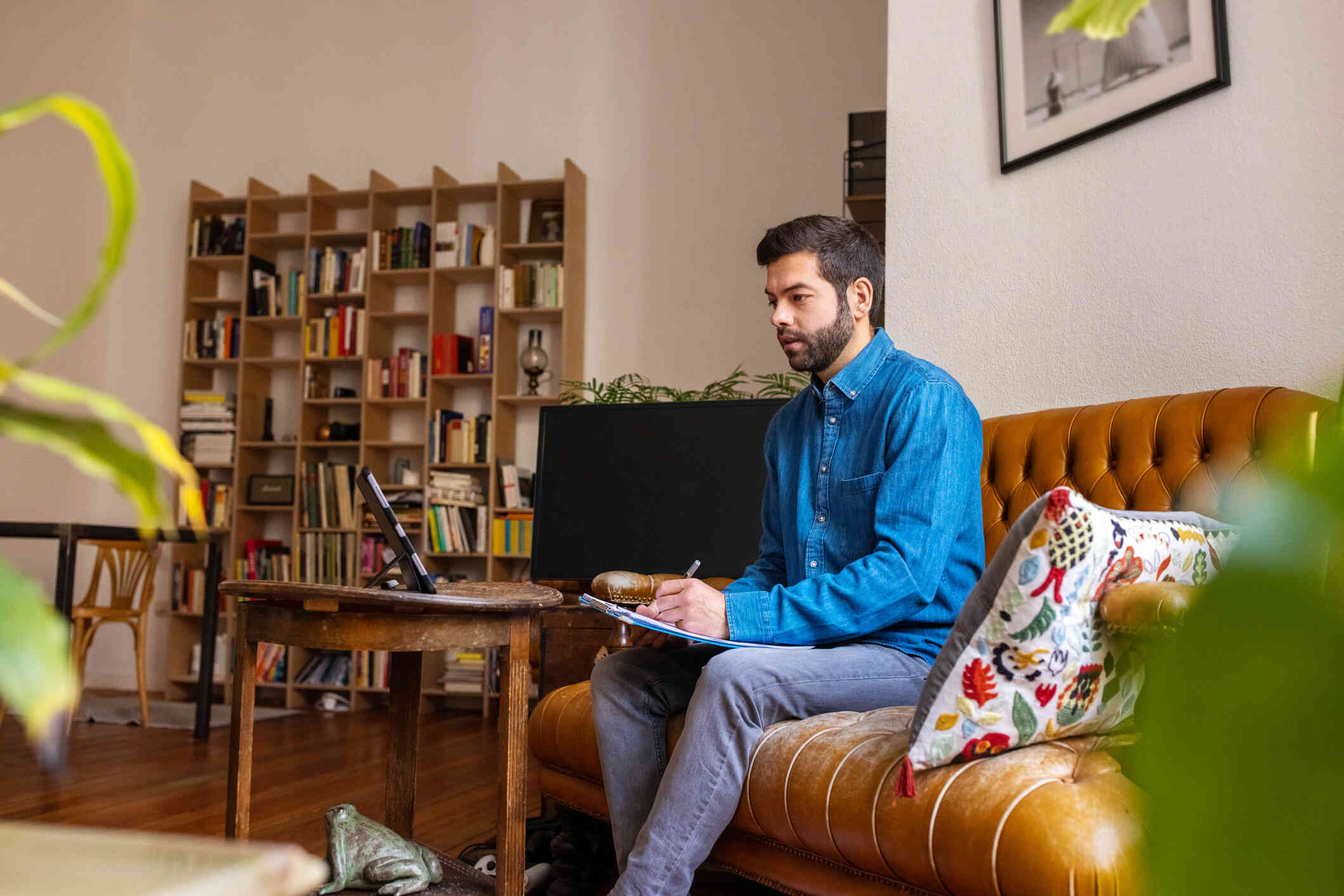 A man in a blue button down shirts sits in his couch while taking notes and looking at a tablet on a table infront of him.