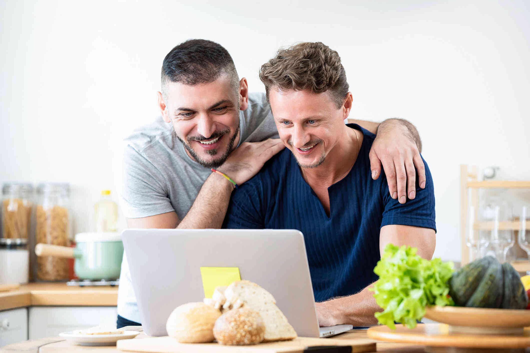 A male couple are at the kitchen table as they both look at the lpatop open infront of them with smiles.