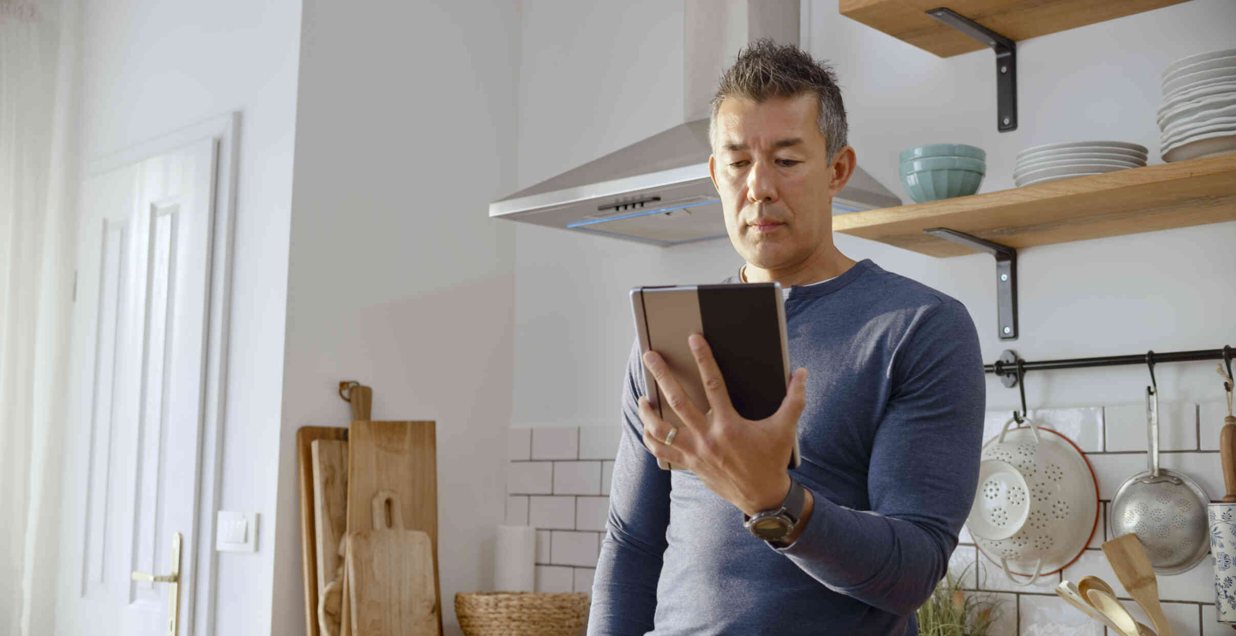 A man with a serious expresson wears a blue sweater as he stands in the kitchen holding a tablet and reading off the screen.