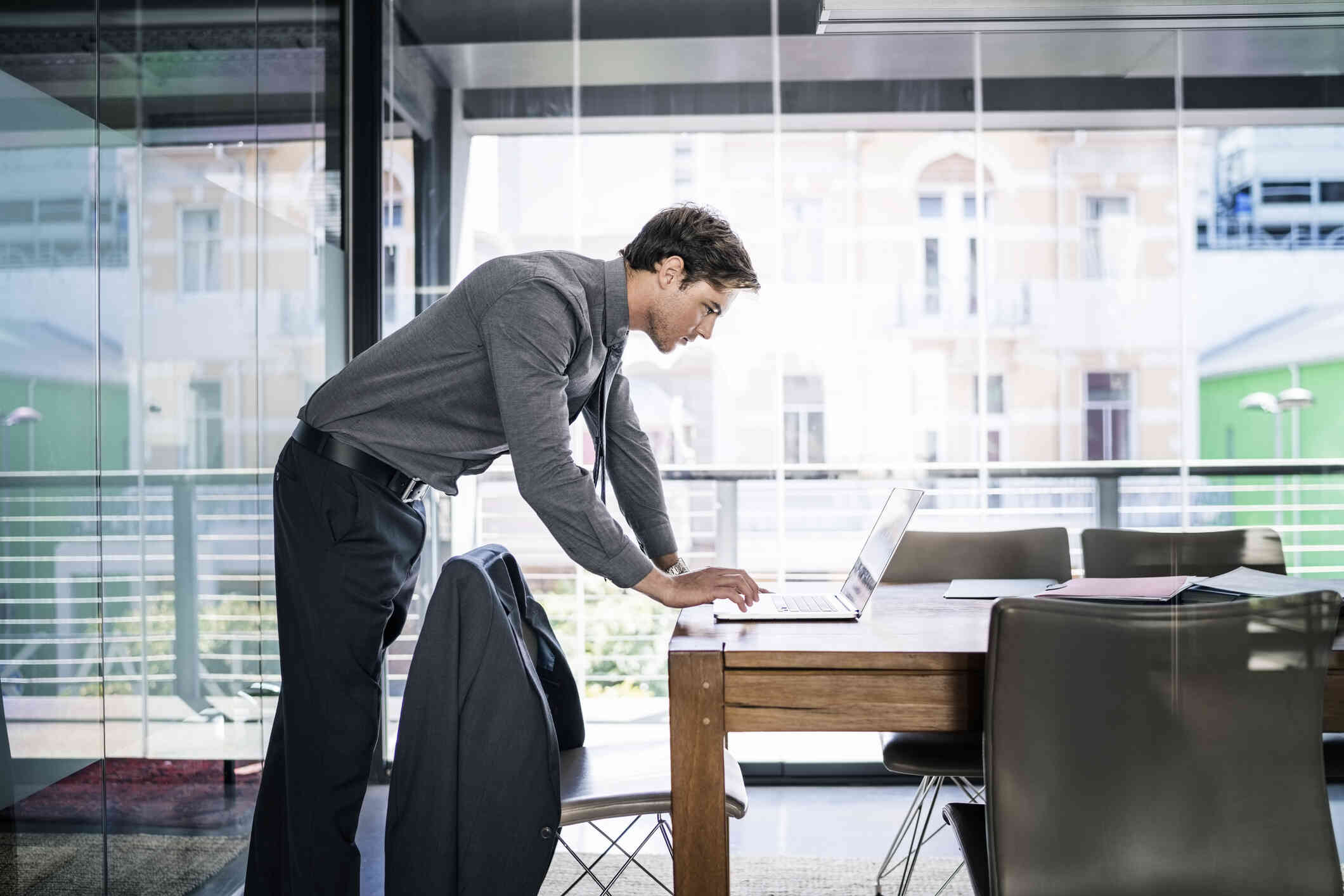 A man in a grey button down shirt stands in a cofference room and and leans down to look at the laptop open on the table.