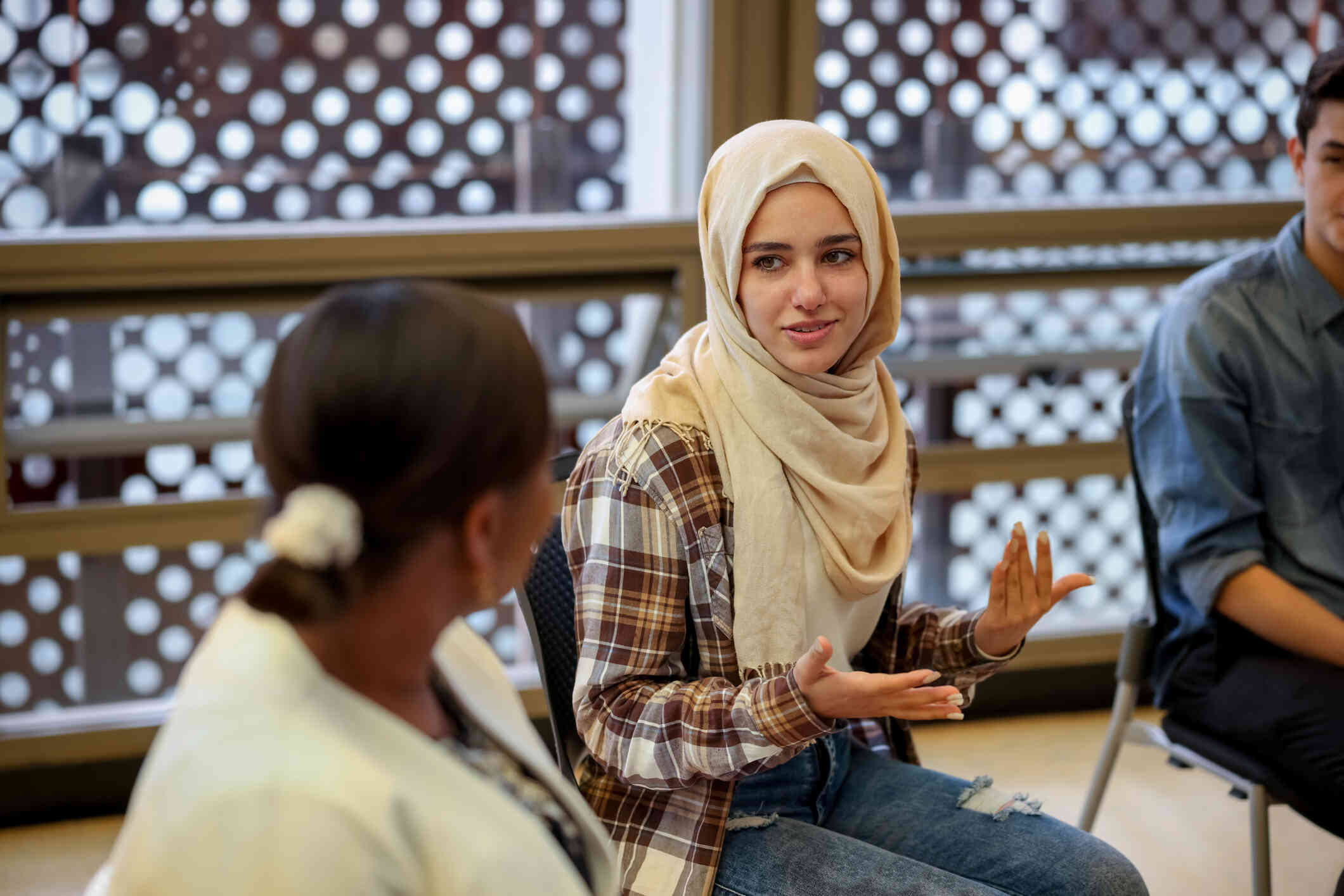 A woman wearing a tan hijab sits in a curcle with other adults and talks during a group therapy session.