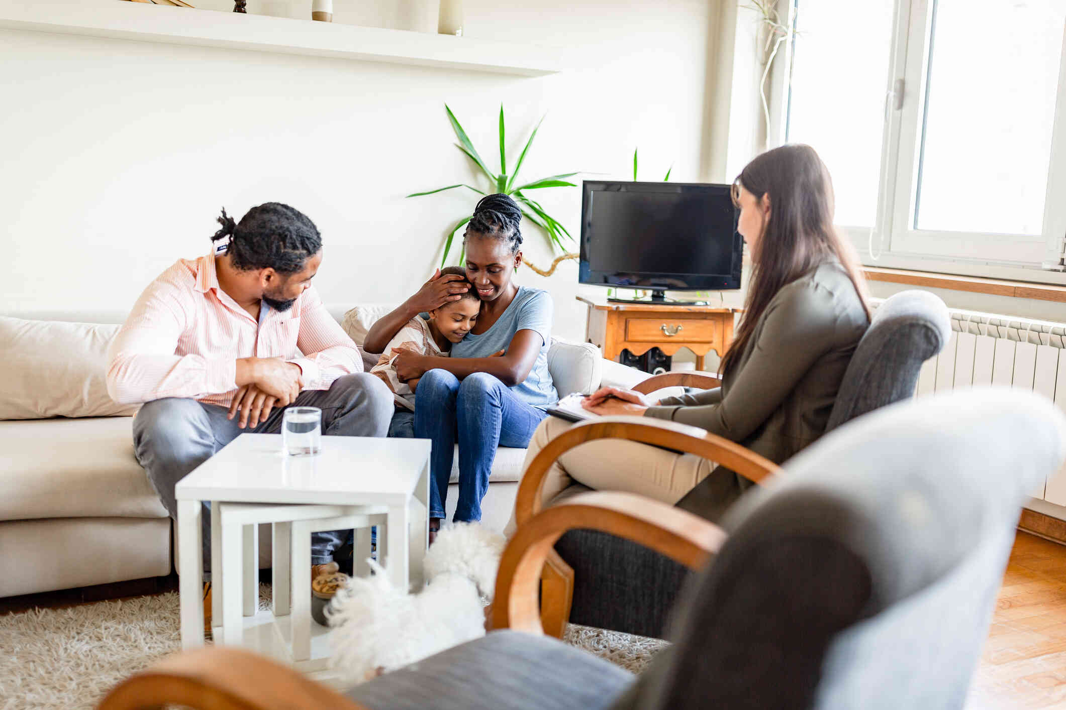 A mother sits on a couch next to her husband while hugging her young son as a female therapist sits across from them during a family therapy session.