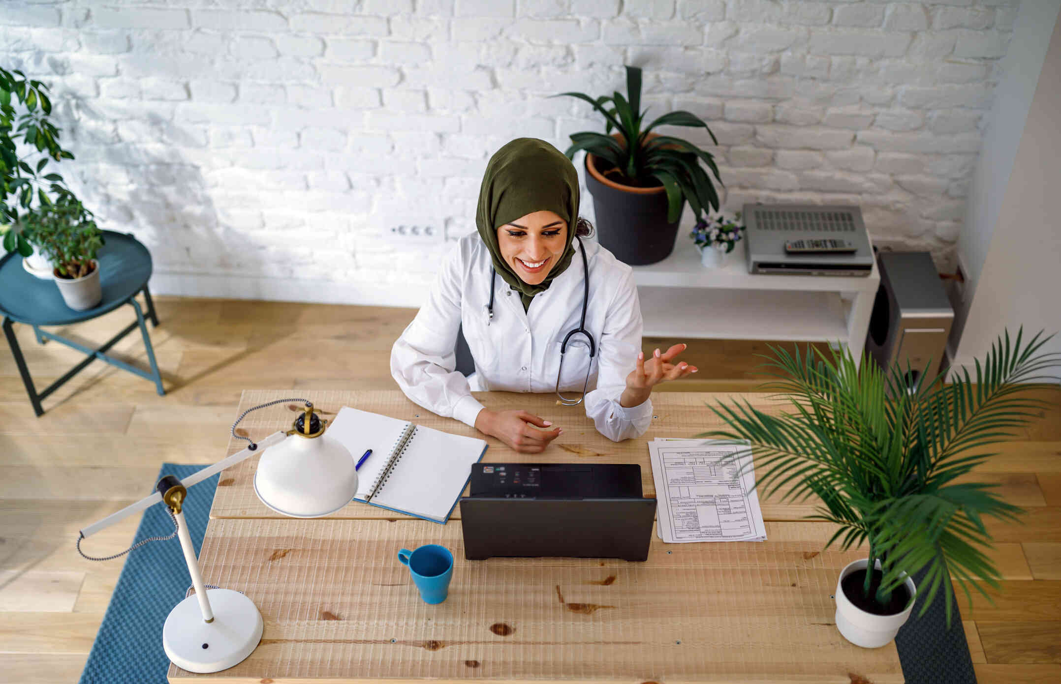 A woman in a doctors coat sits at a wooden table and talk to her laptop screen with a smile during a telehealth call.