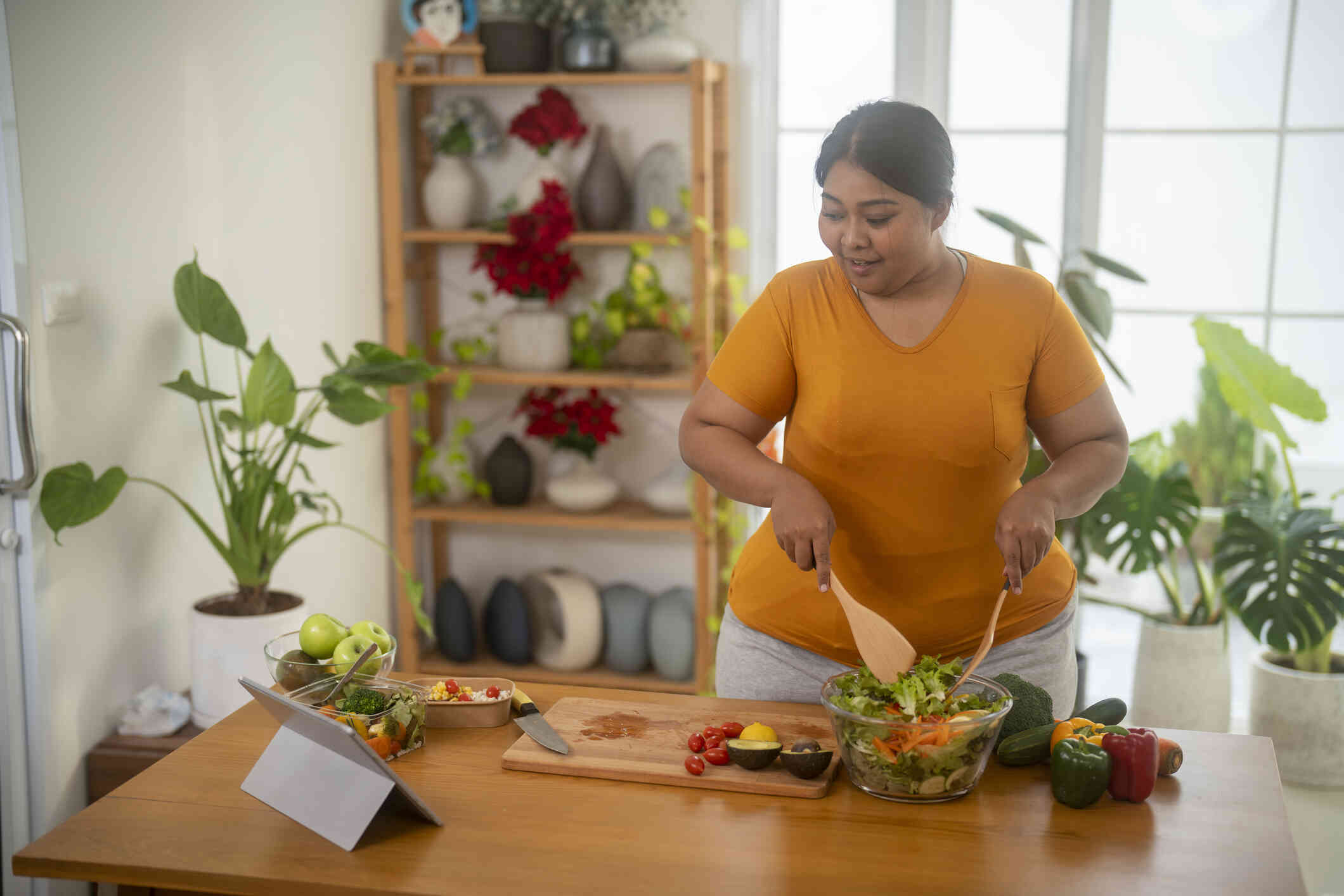 A woman in an ornage shirt sitands in her kitchen while cooking as she looks at the tablet proped up on the counter infront of her.