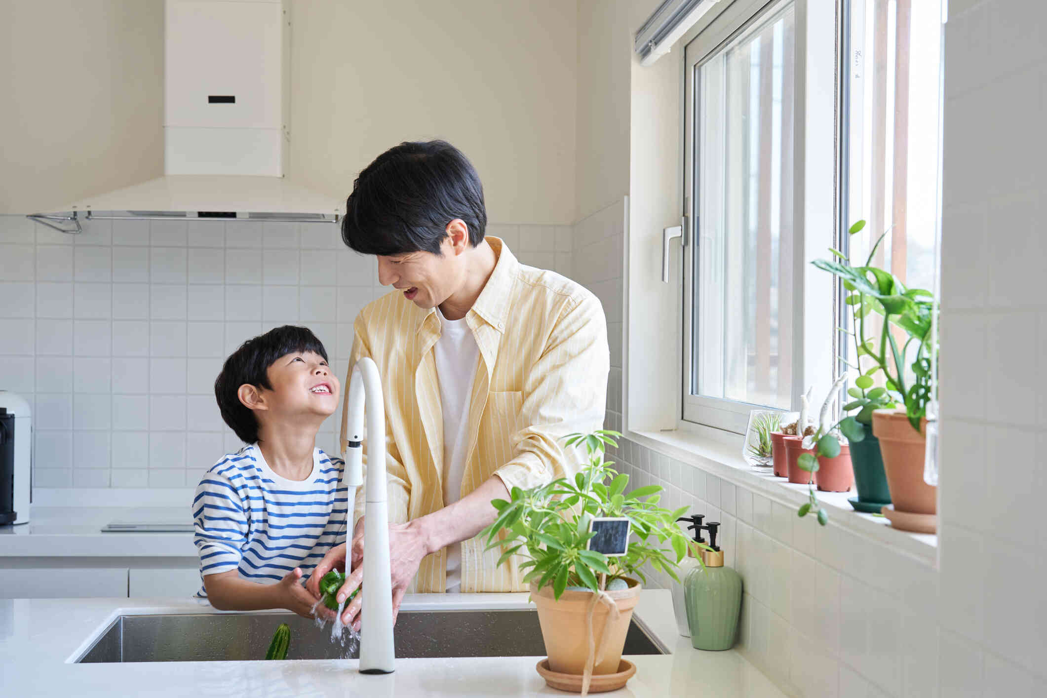 A man and his young son stands next to each other at the kitchen sink while washing peppers and smiling at one another.