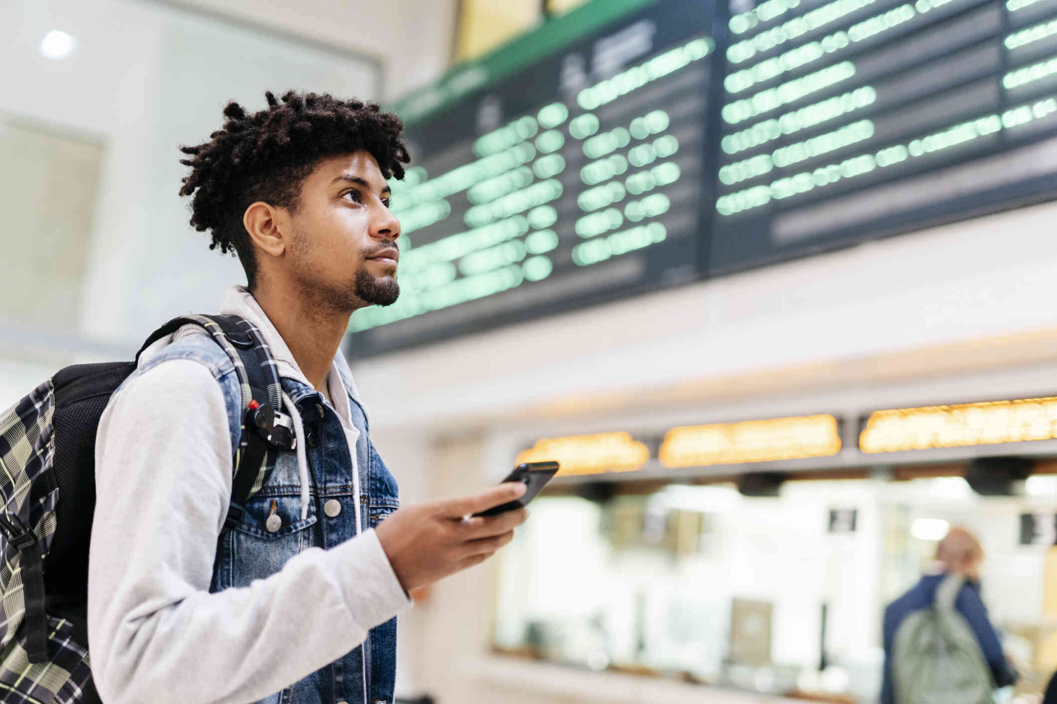 A man wearing a backpack stands in an airport and looks around as he holds a cell phone in his hand.