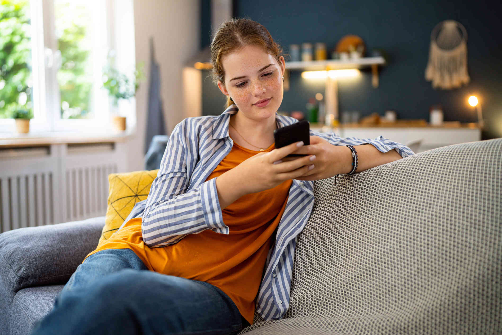 A teen girl in an orange shirt sits casually on the couch in the living room and looks at the cellphone in her hand.