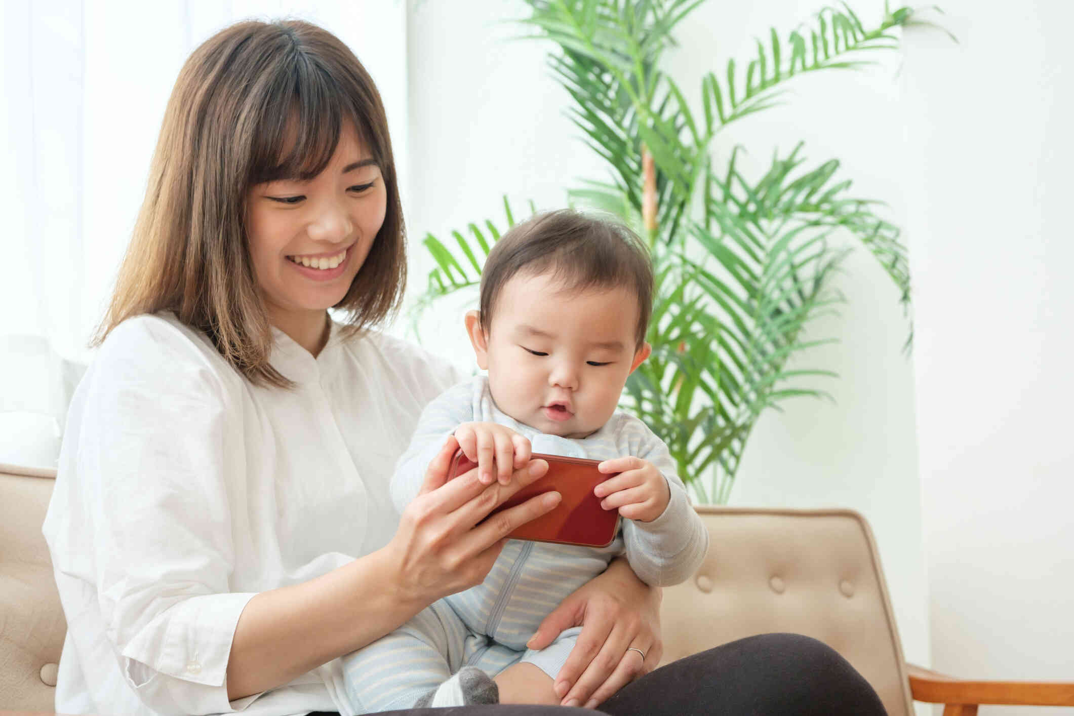 A mother in a white shirt sits in her home and holds her inafant child and her phone while smiling.