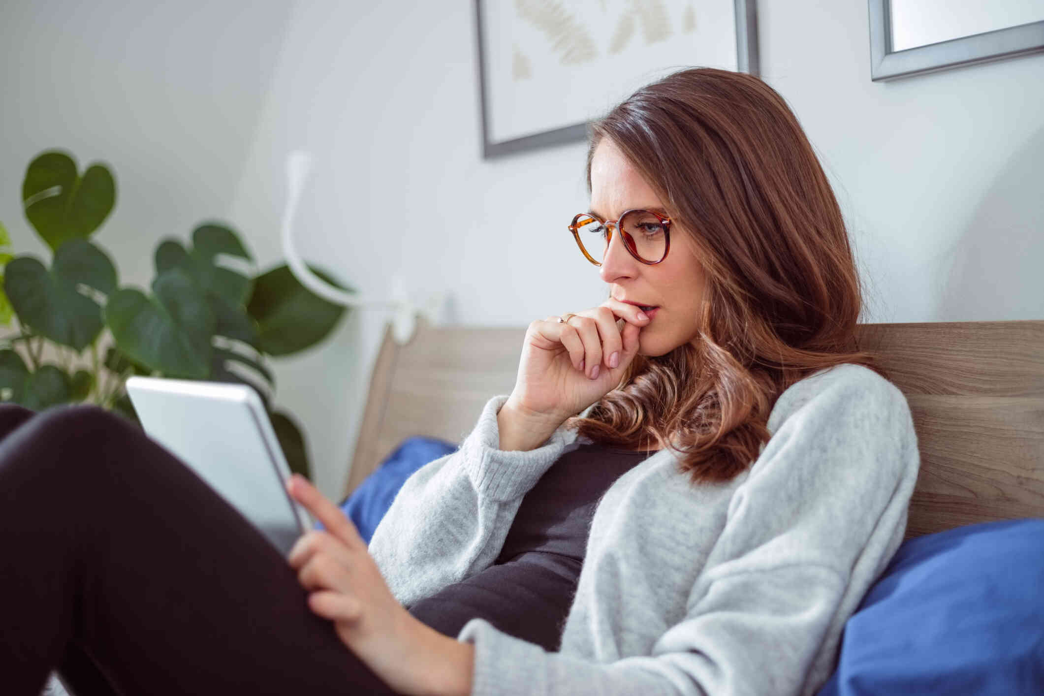 A woman with glaes sits up in bed with her back against the the headboard as she looks at the tablet against her legs.