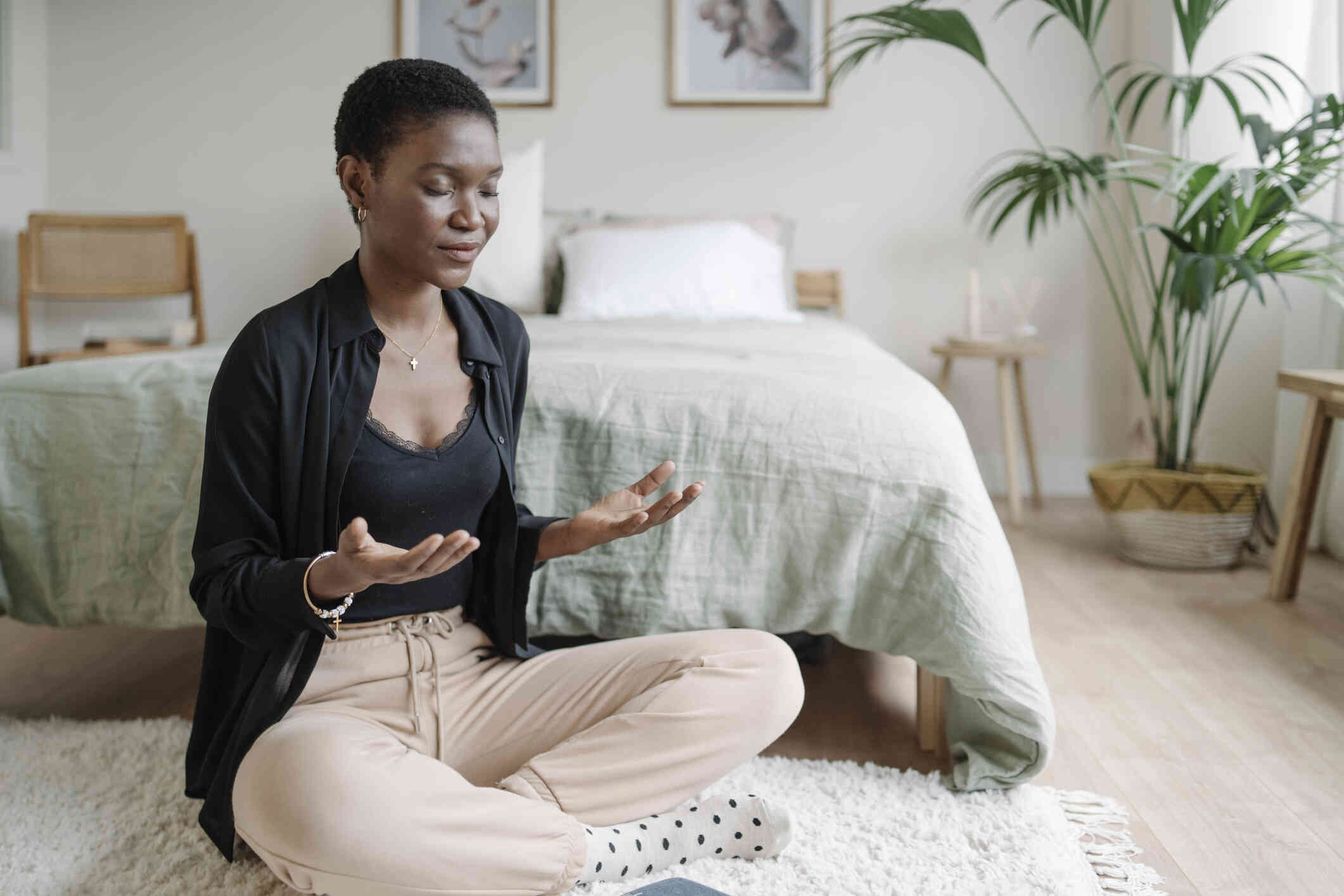 A woman with a calm expression and closed eyes sits at the foot of her bed and meditates with her legs crossed and her arms raised in front of her.