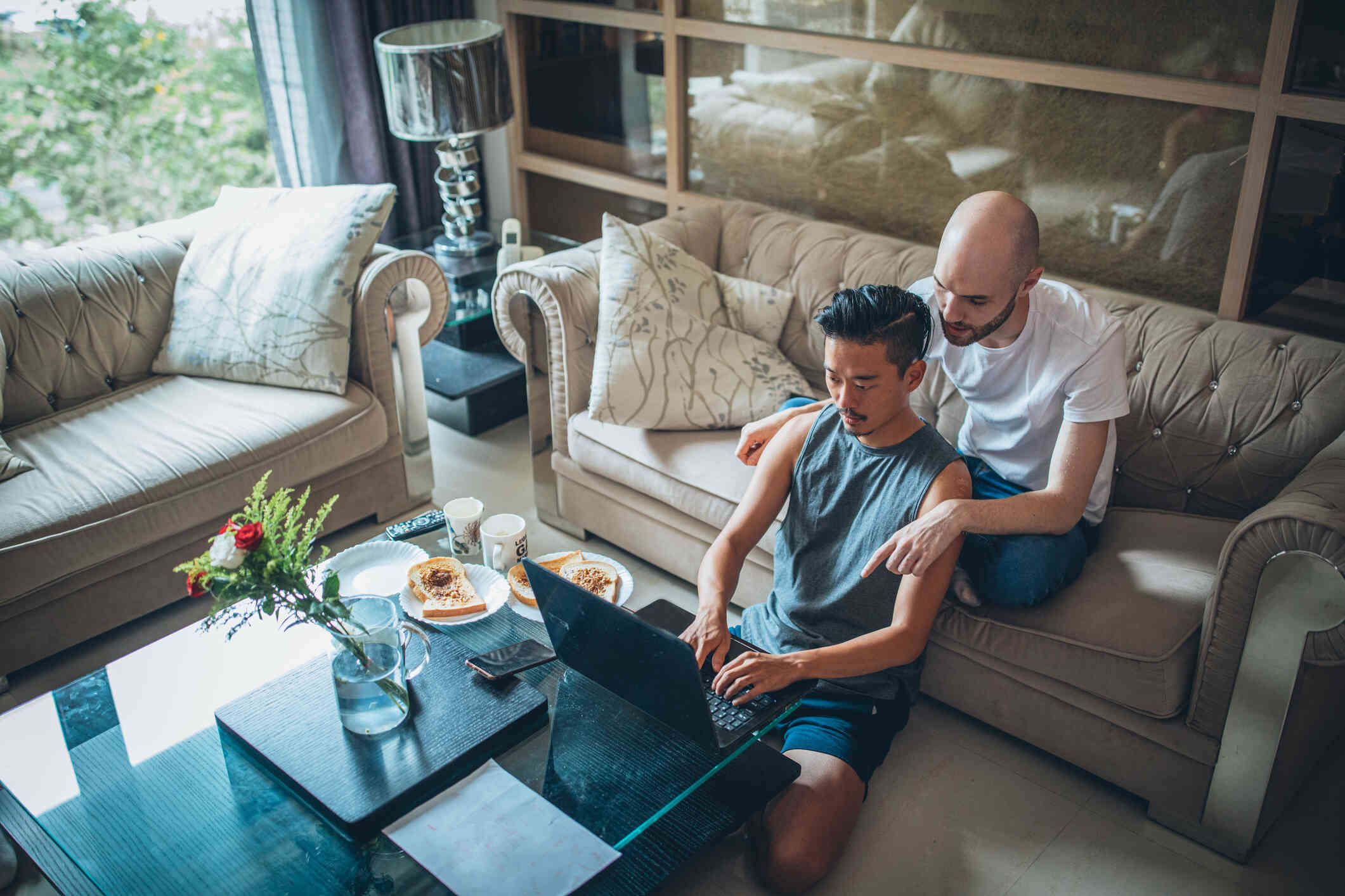 A man sits on the floor while typing on a laptop on the coffee table infront of him as his male partner sits on the couch behind him.