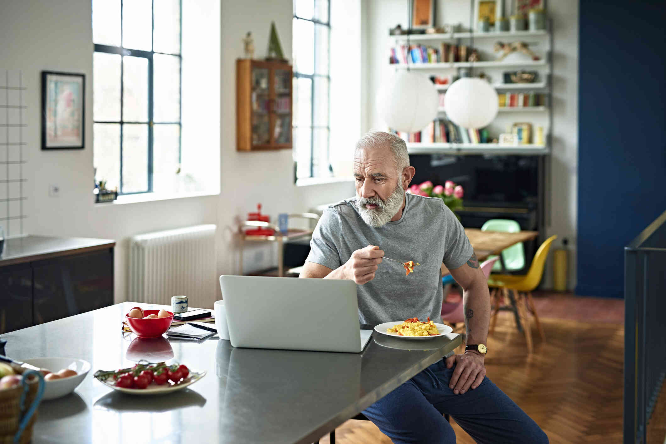 A mature man in a grey shirt sits at the ktichen table while eating breakfast and looking at the laptop open in the table infrot of him with a serious expression.