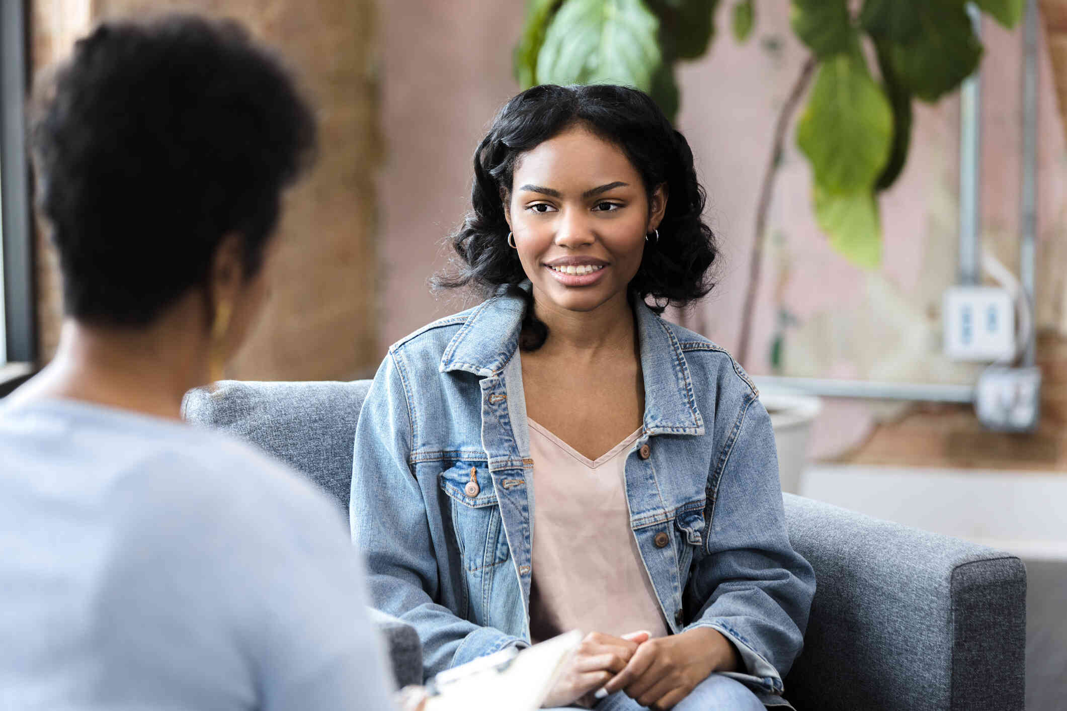 A woman in a blue jean jacket sits across from her female specialist and listens to her talk with a smile.