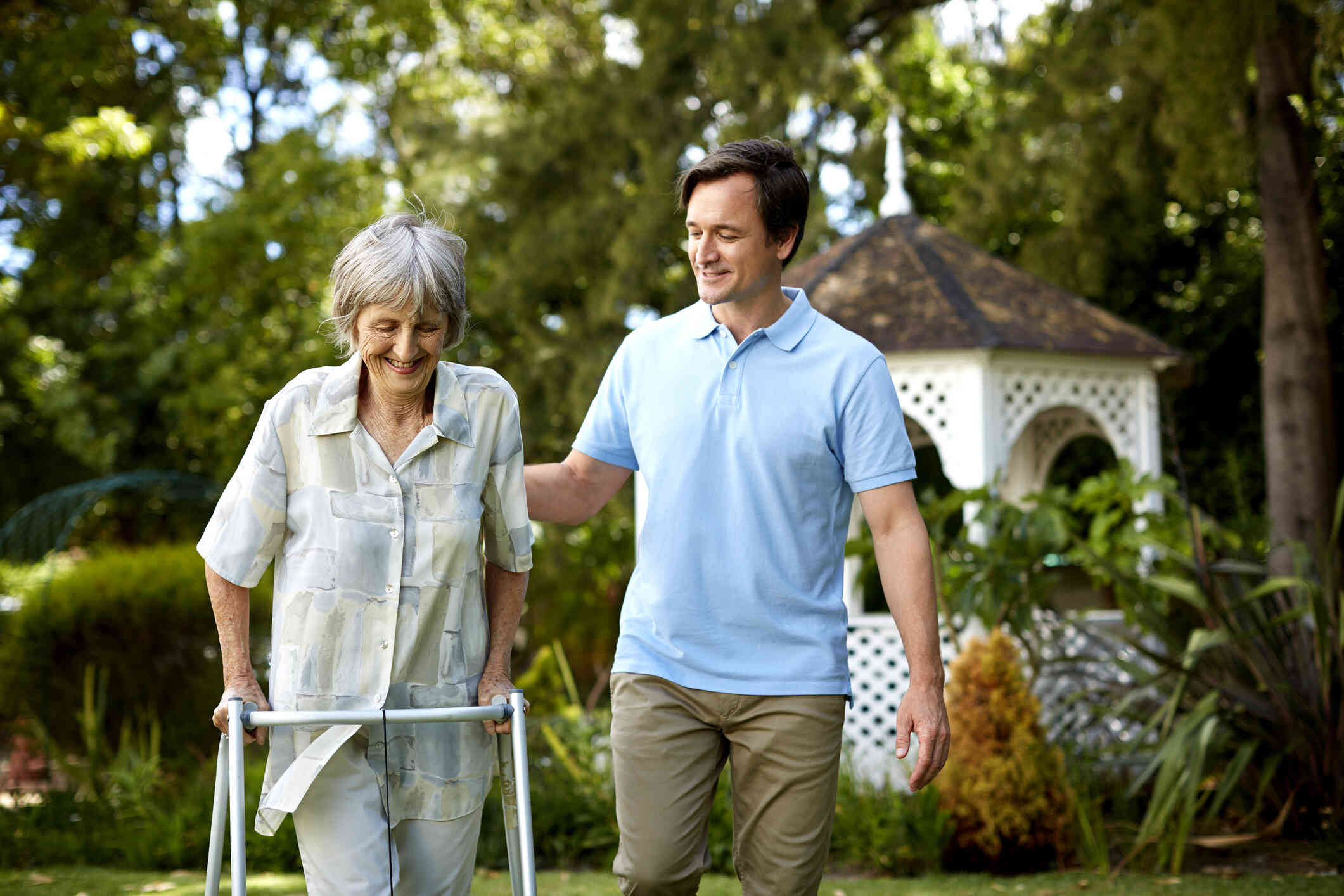 A man in a blue shirt walks alongside his elderly mother with a walker as the smile and go for a walk on a sunny day.