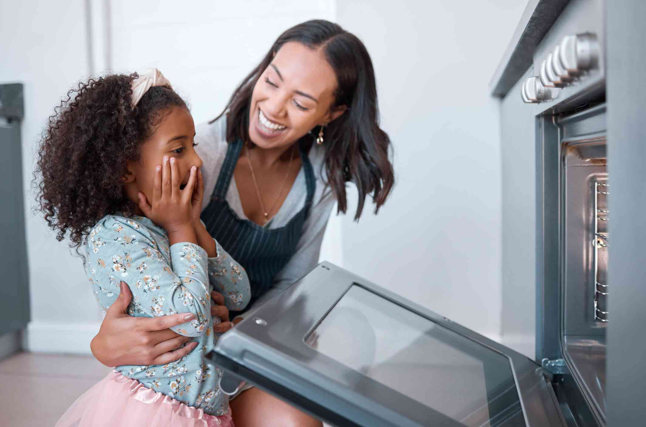 A mother and daughter are in the kitchen in front of an open oven; the mom is looking at the daughter and smiling. 