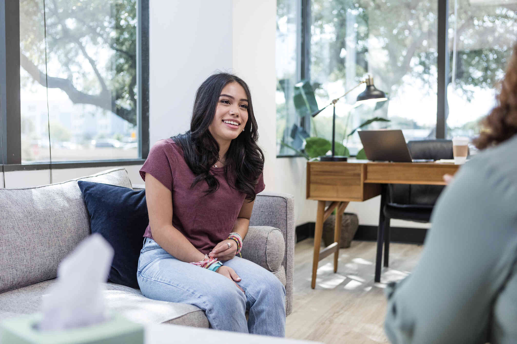 A young woman in a maroon shirt with a positive expression sits on a grey couch while engaged in a conversation with a counselor