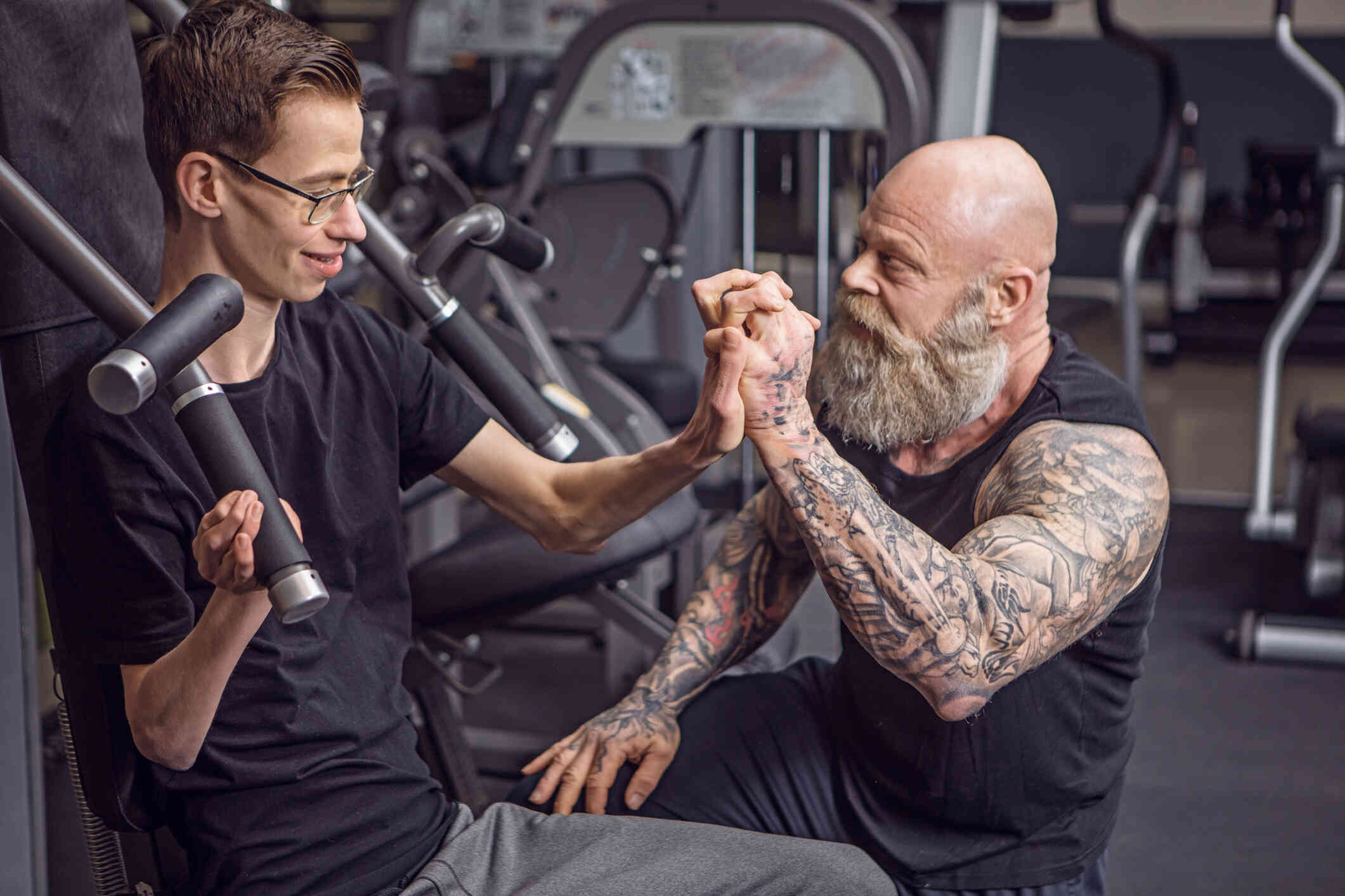 A man with glasses sits on a piece of exercise equipment and lifts weights while clasping the hand of another man with tatoos on his arms who sits next to him with an encouraging expression.