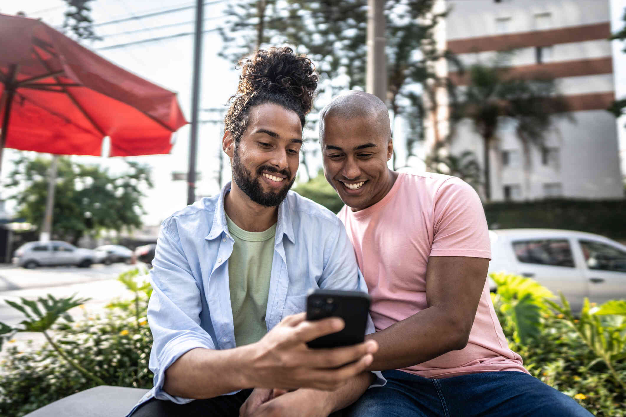 A male couple sit next to each outher outside on a sunny day while looking at a phone together and smiling.