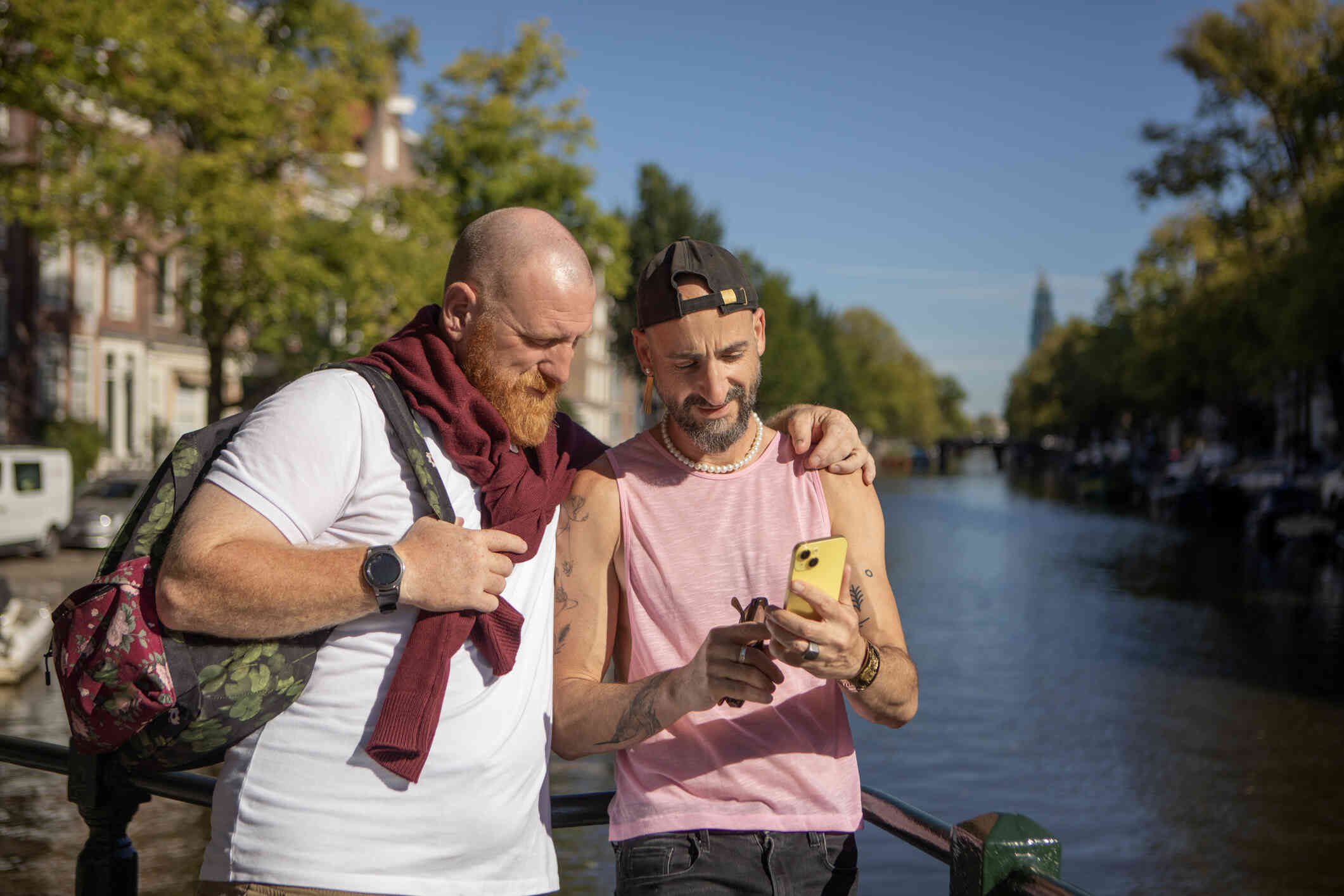 A male couple stand outside on a sunny day while looking at a phone screen together.