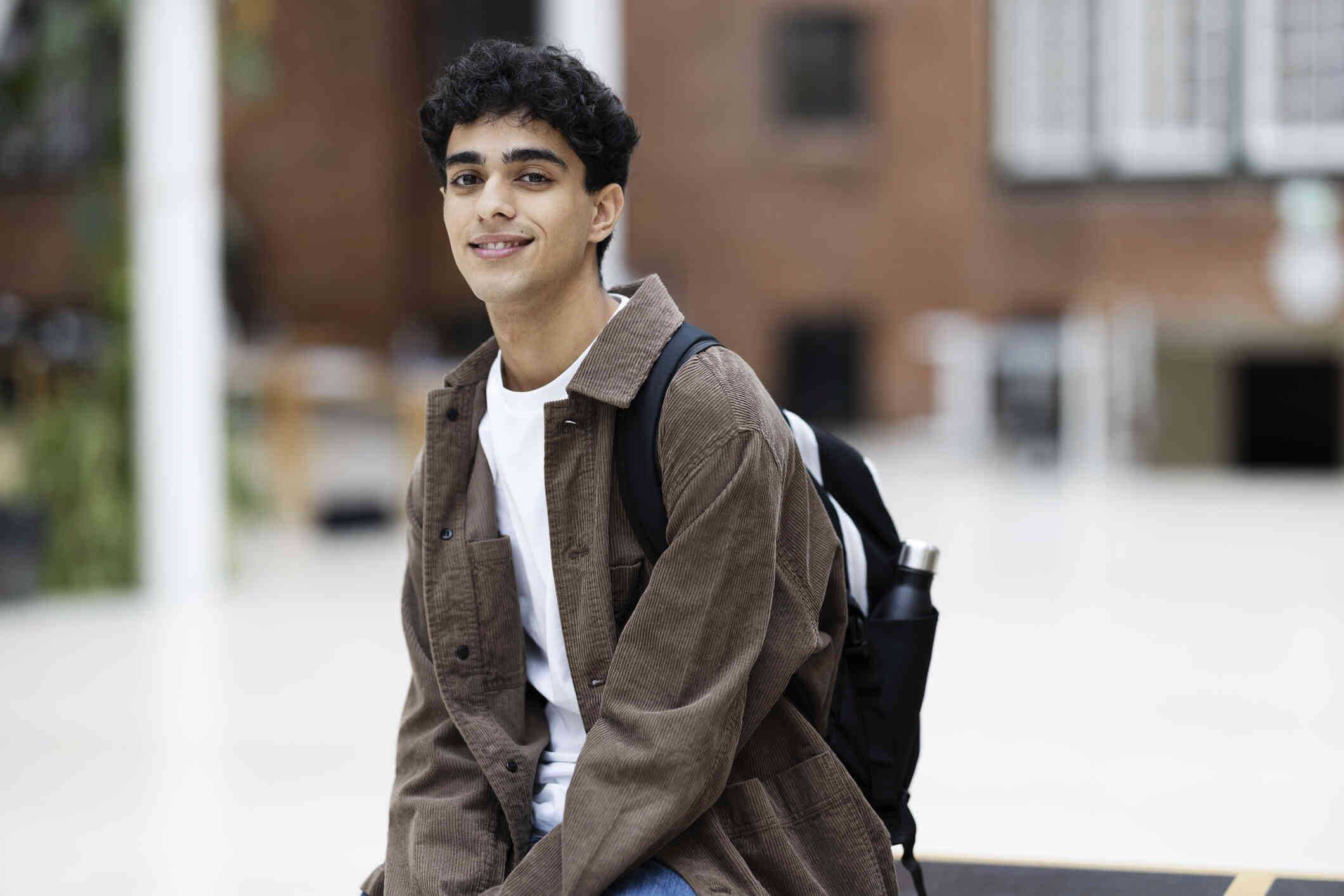 A young man with dark hair and a brown jacket wears a backpack as he sits in front of a college building smiling.