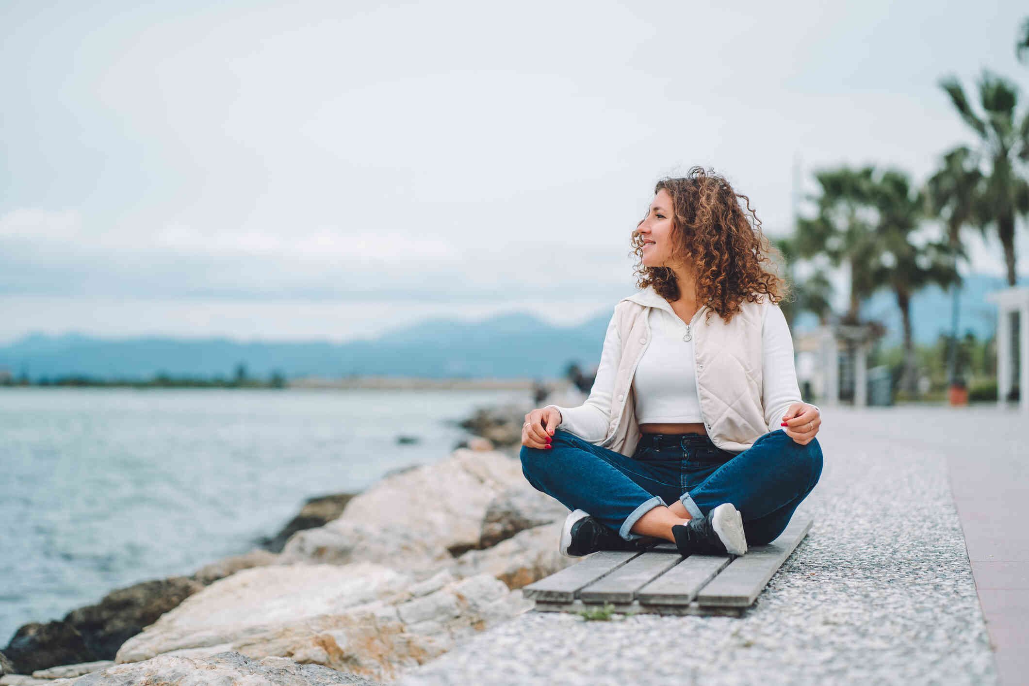 A woman with curly hair and a content expression sits outside with her legs crossed and looks out over a body of water next to her.