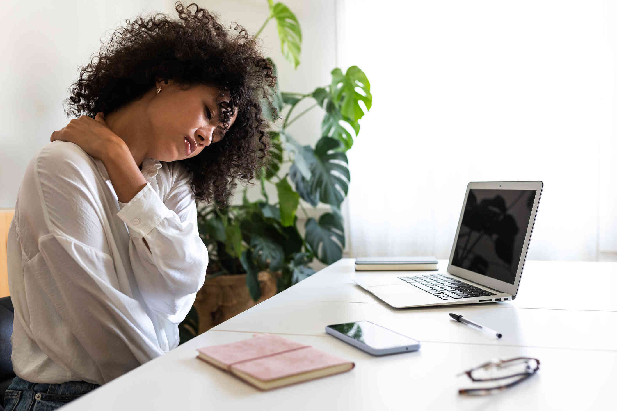 A woman rubs her sore neck while closing her eyes as she sits at her desk with her laptop open infront of her.