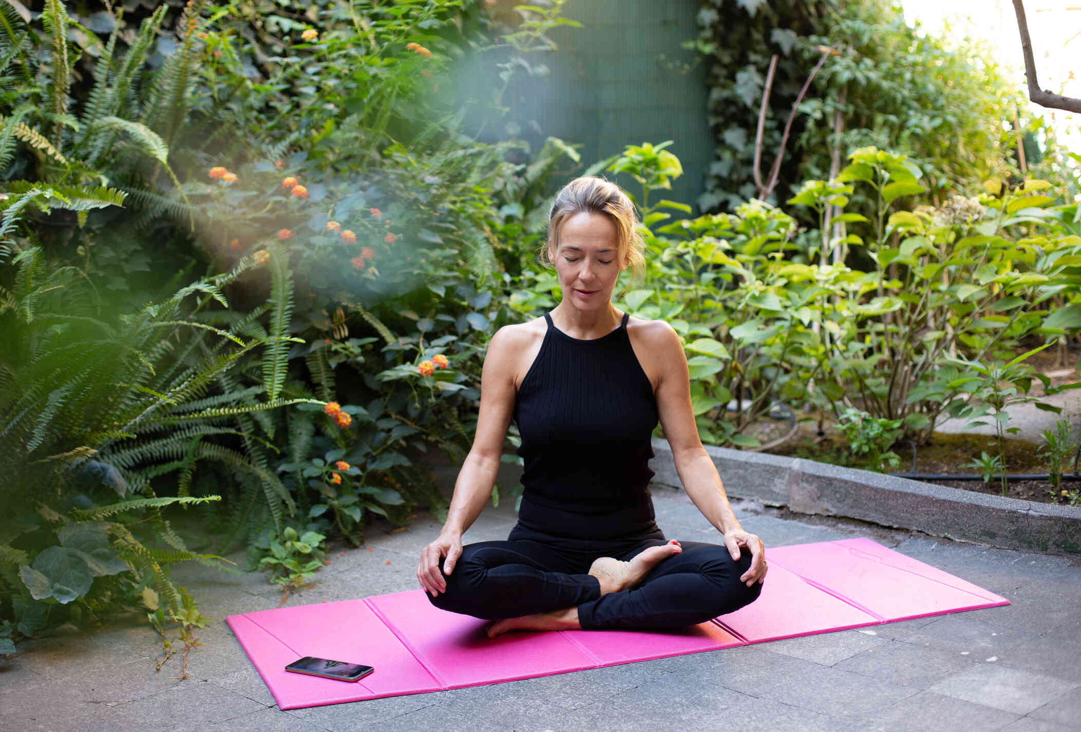 A woman in a black shirtsits on a pink yoga mat outside with her eyes closed.