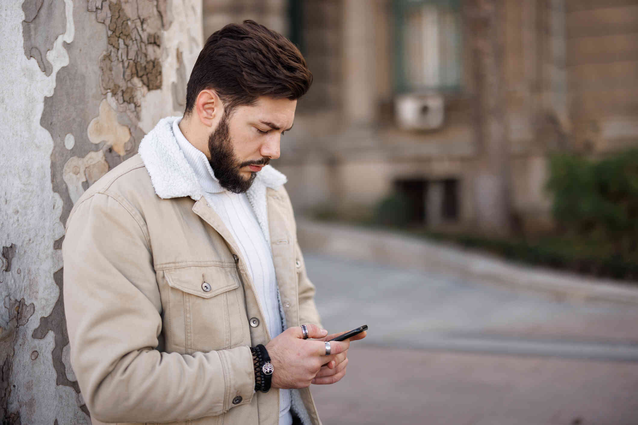 A man in a tan jacket stands outside near a cement wall and looks down at the cellphone in his hands with a serious expression.